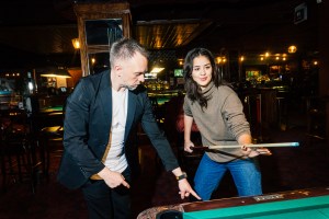 A man in a blazer and a white t-shirt points at a pool table, a woman stands next to him holding a cue stick.