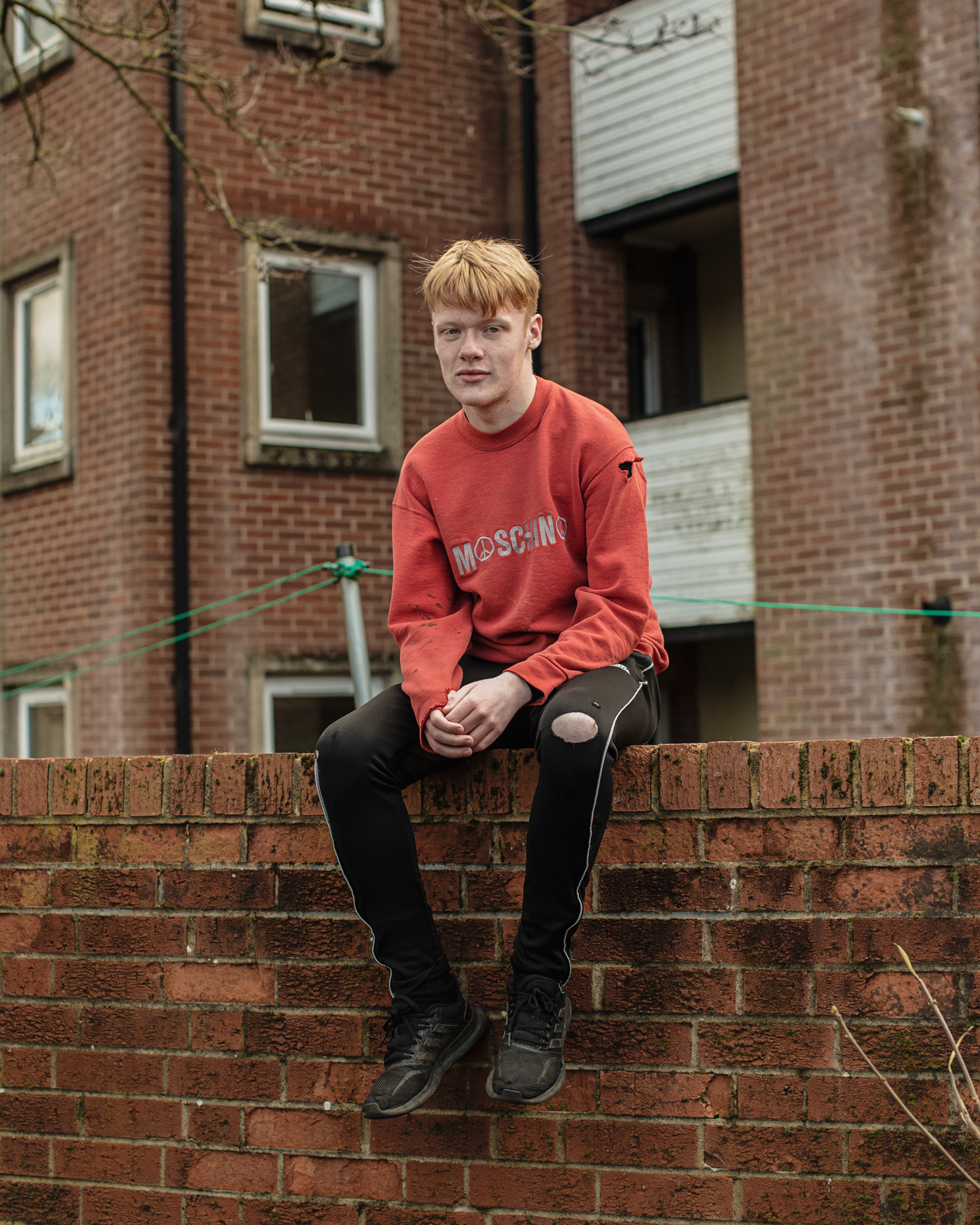 A young man in a ripped sweater and track pants sits on a brick wall.