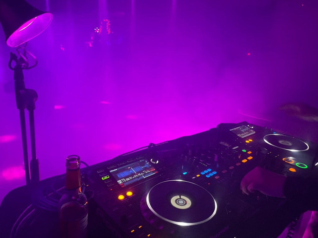 A photo from behind DJ decks looking out into a dancefloor filled with hazy purple lights