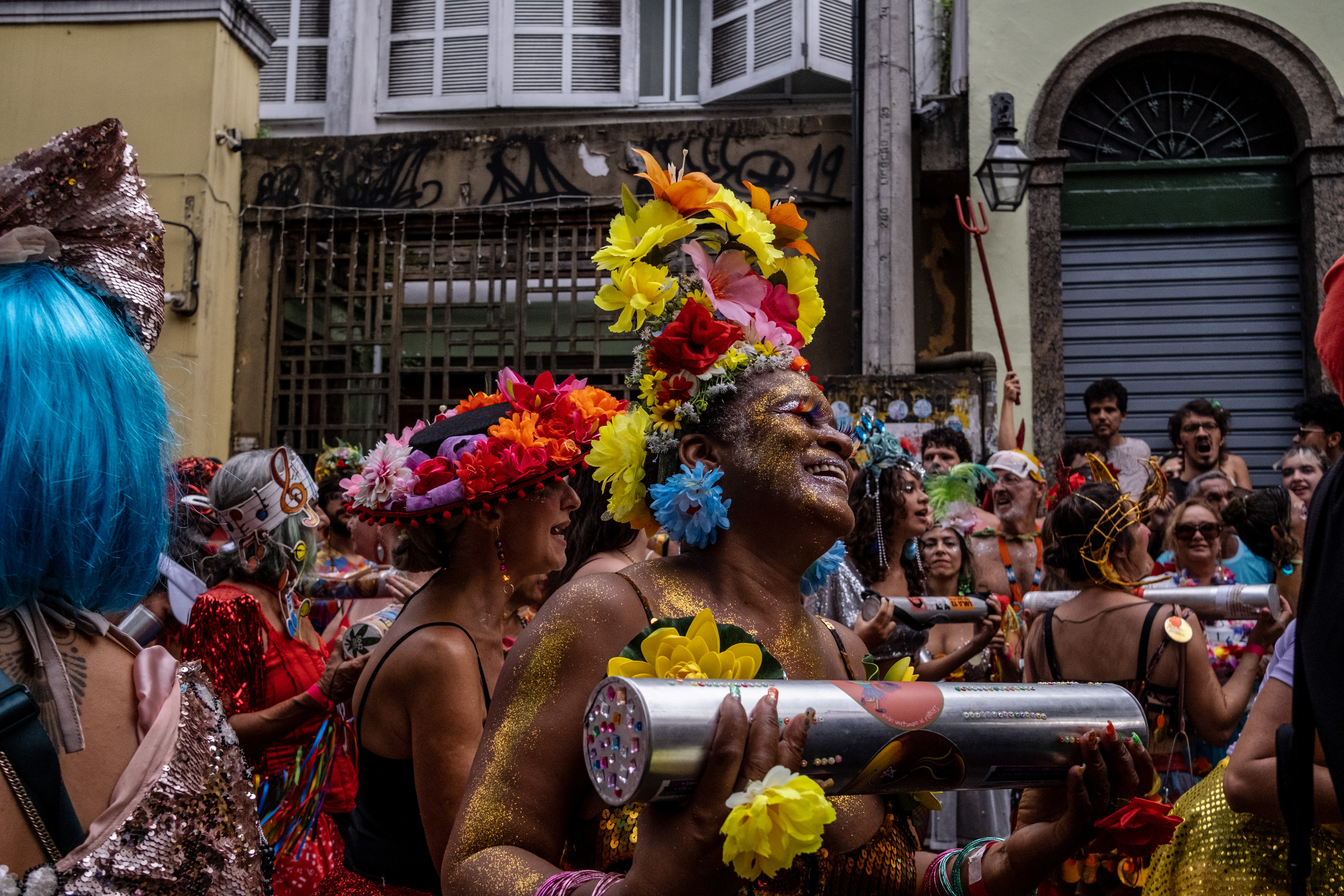 A person covered in glitter and wearing a colorful floral headpiece holds a shaker instrument in a crowded street of performers.