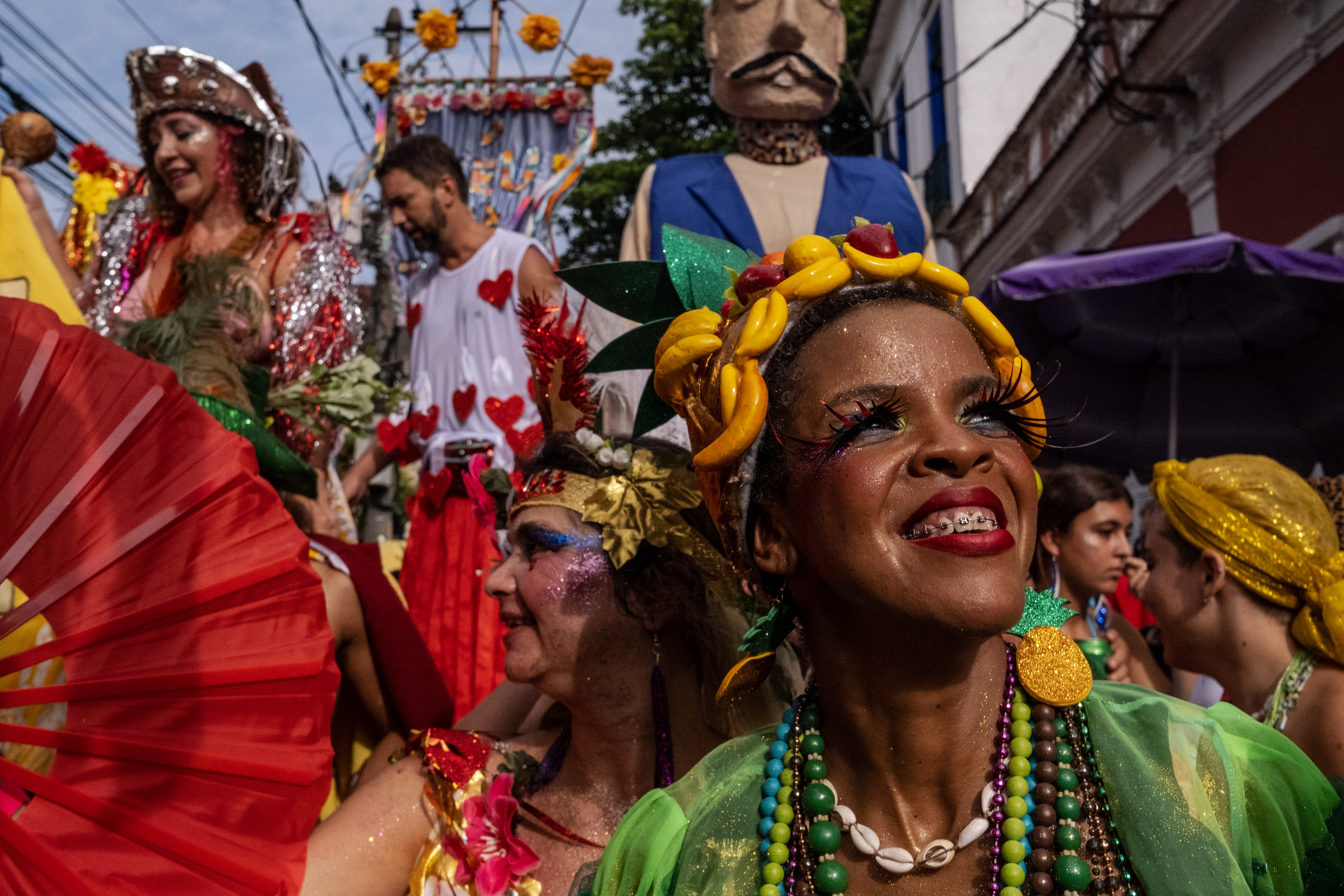 A woman with colorful beads, very long decorative lashes, and braces smiles as she looks into the sun. Behind her is a packed street of performers.