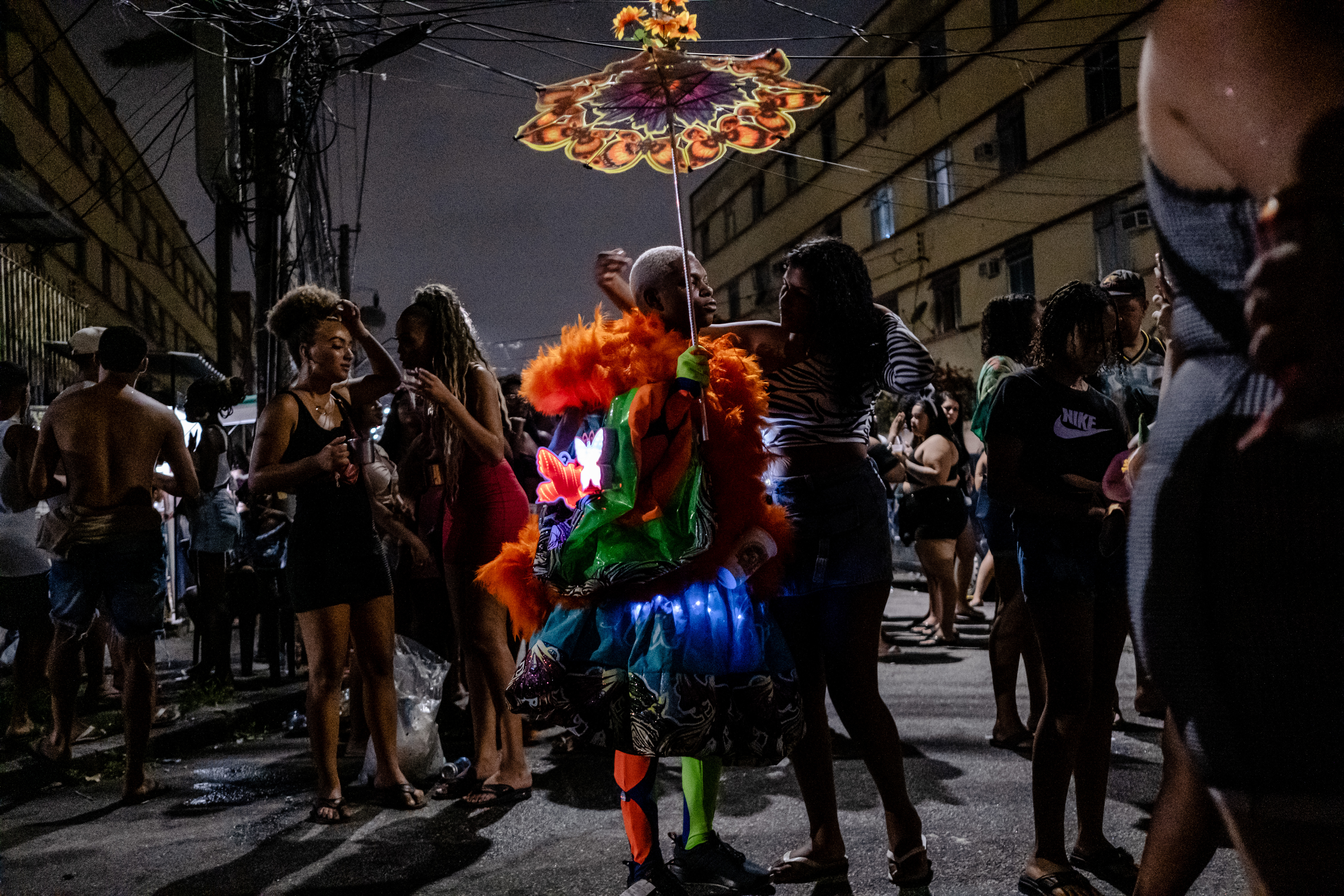 A person in a colorful traditional costume holds a decorative umbrella while someone else embraces them from the side. It's nighttime and the streets are more sparse.