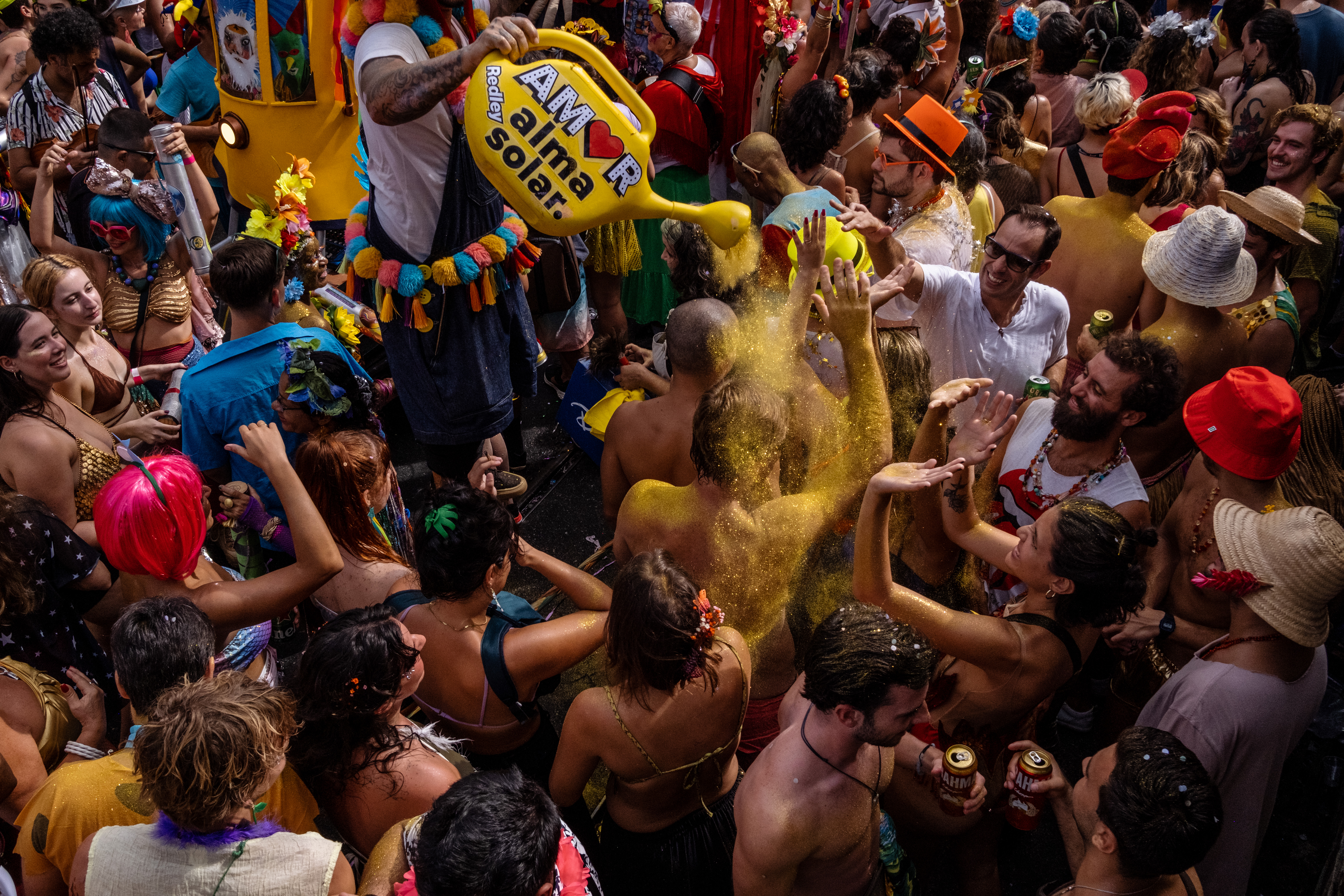 A person on stilts pours yellow pigment out of a plastic watering can on partygoers. The street is completely crowded with people.