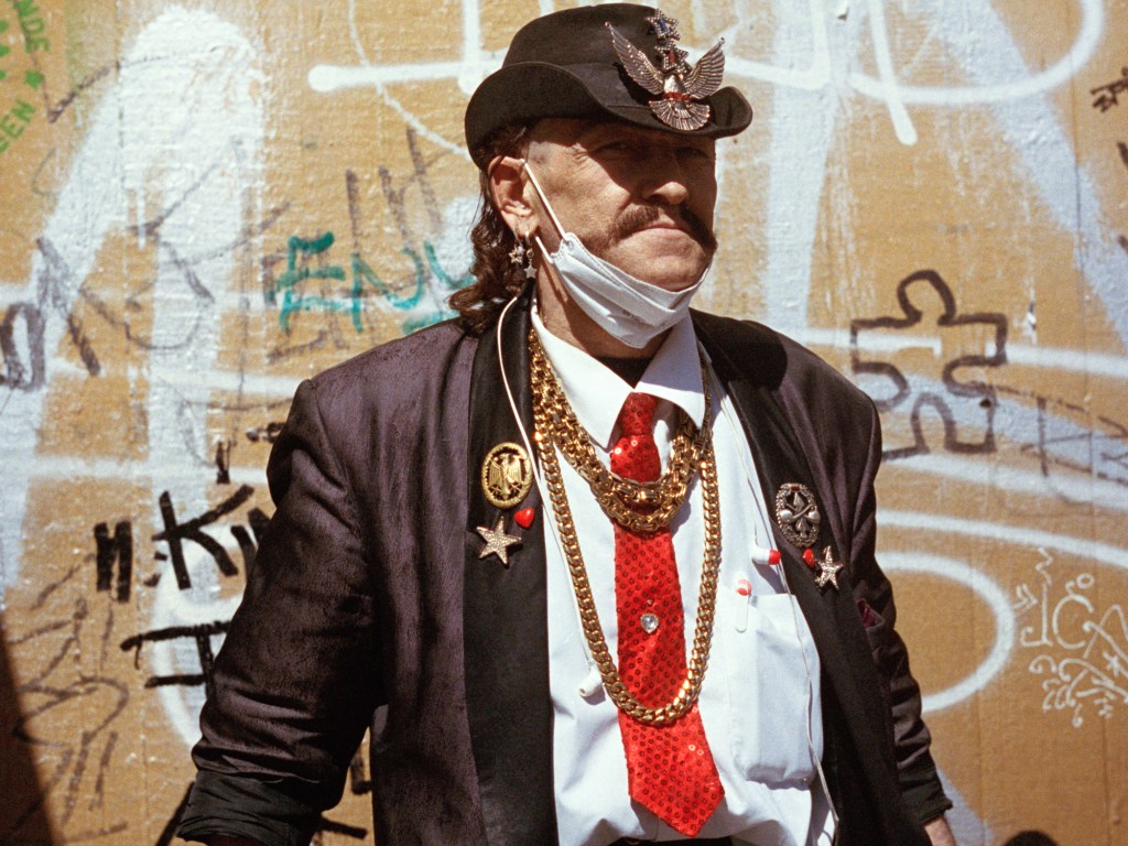 A man wearing a brown cowboy hat and jacket with golden decorations. He also wears a white shirt and a red tie as well as golden necklaces. He has long hair.