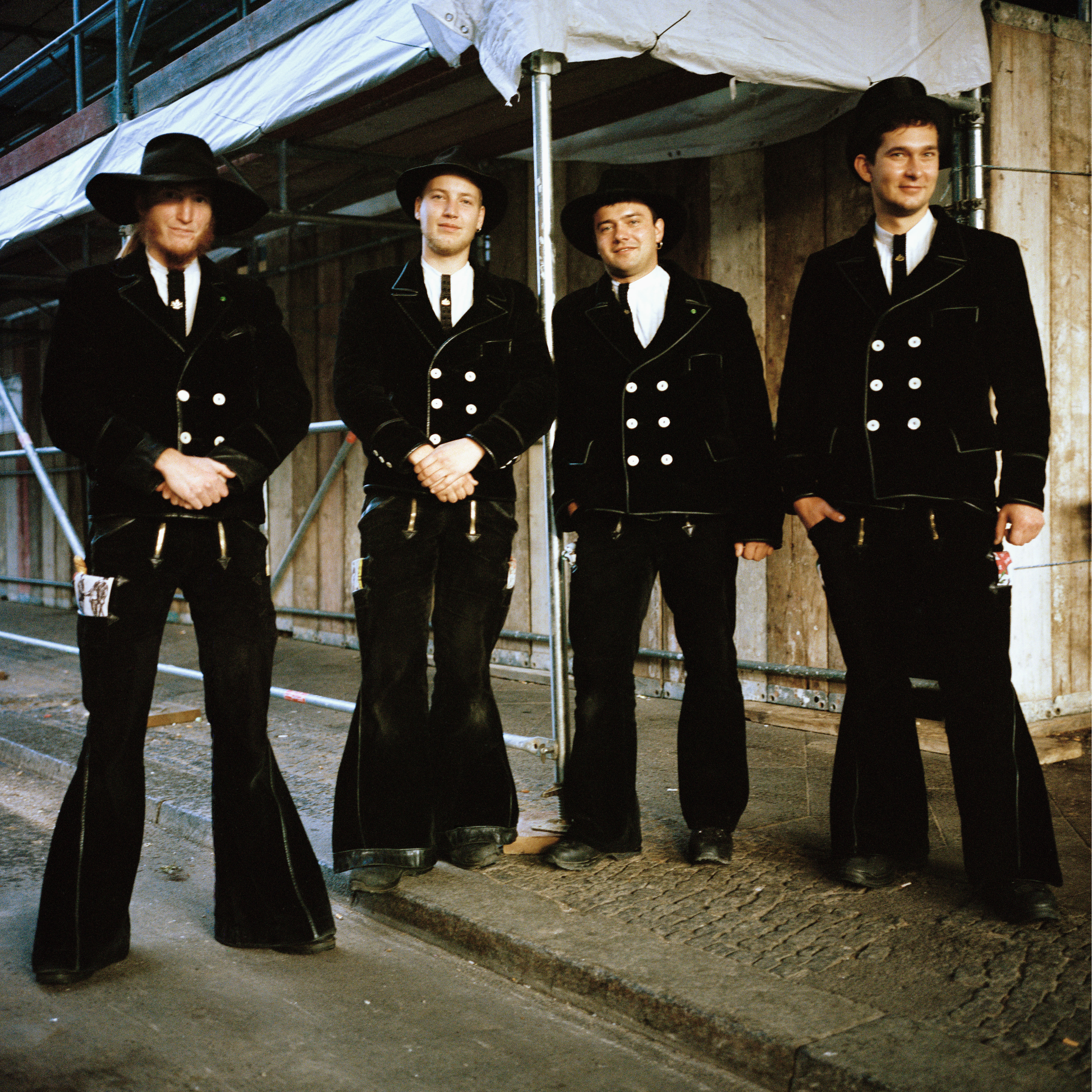 Kottbusser Tor, Berlin. Four men wearing a black costume with a hat. In the background there's a façade under renovation.