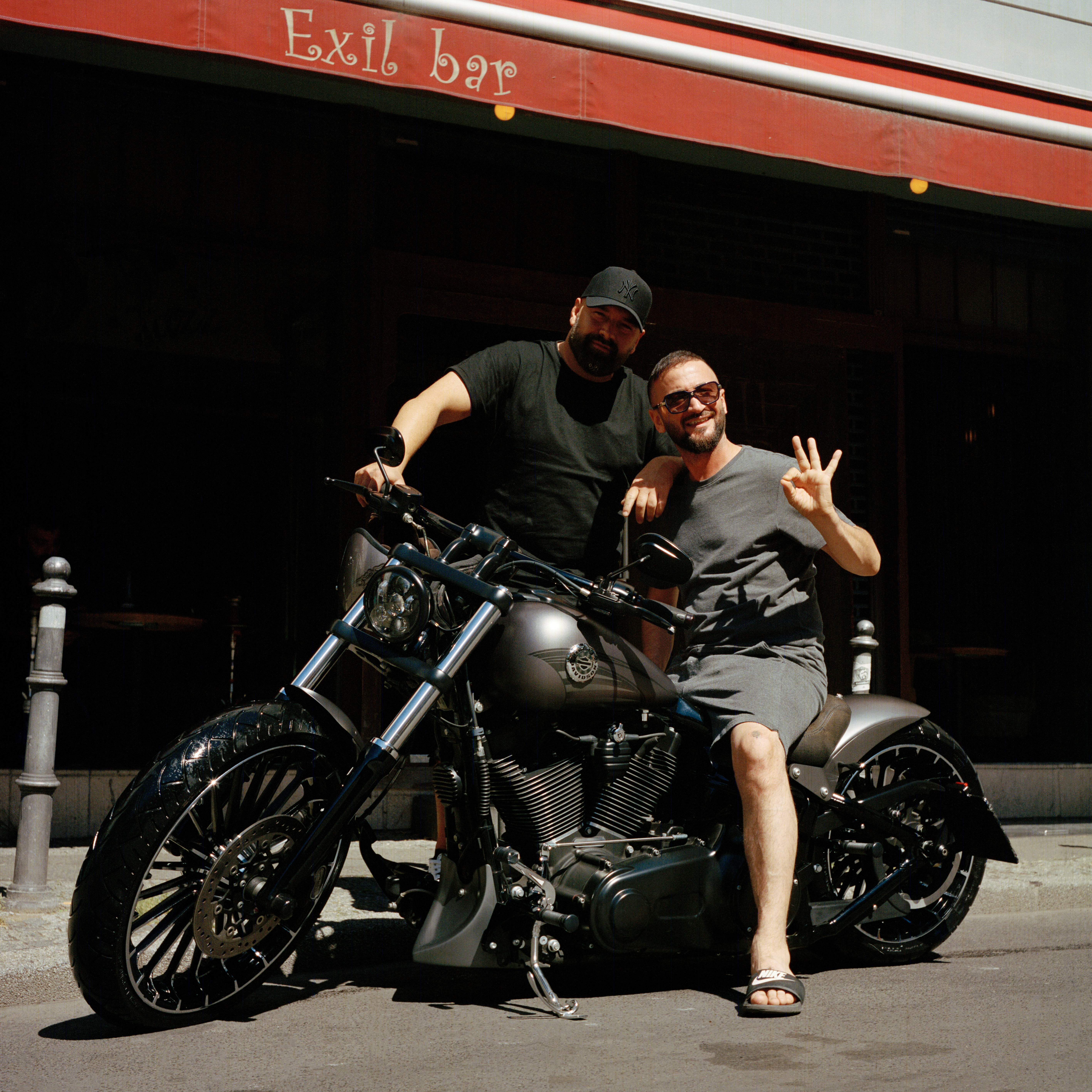 Kottbusser Tor, Berlin. Two bearded dark-haired men dressed in black in front of Exil Bar in the summer: one of them sits on a big, black, shiny motorbike and wears shorts, a t-shirt and flipflops. The other one stands behind the motorbike.