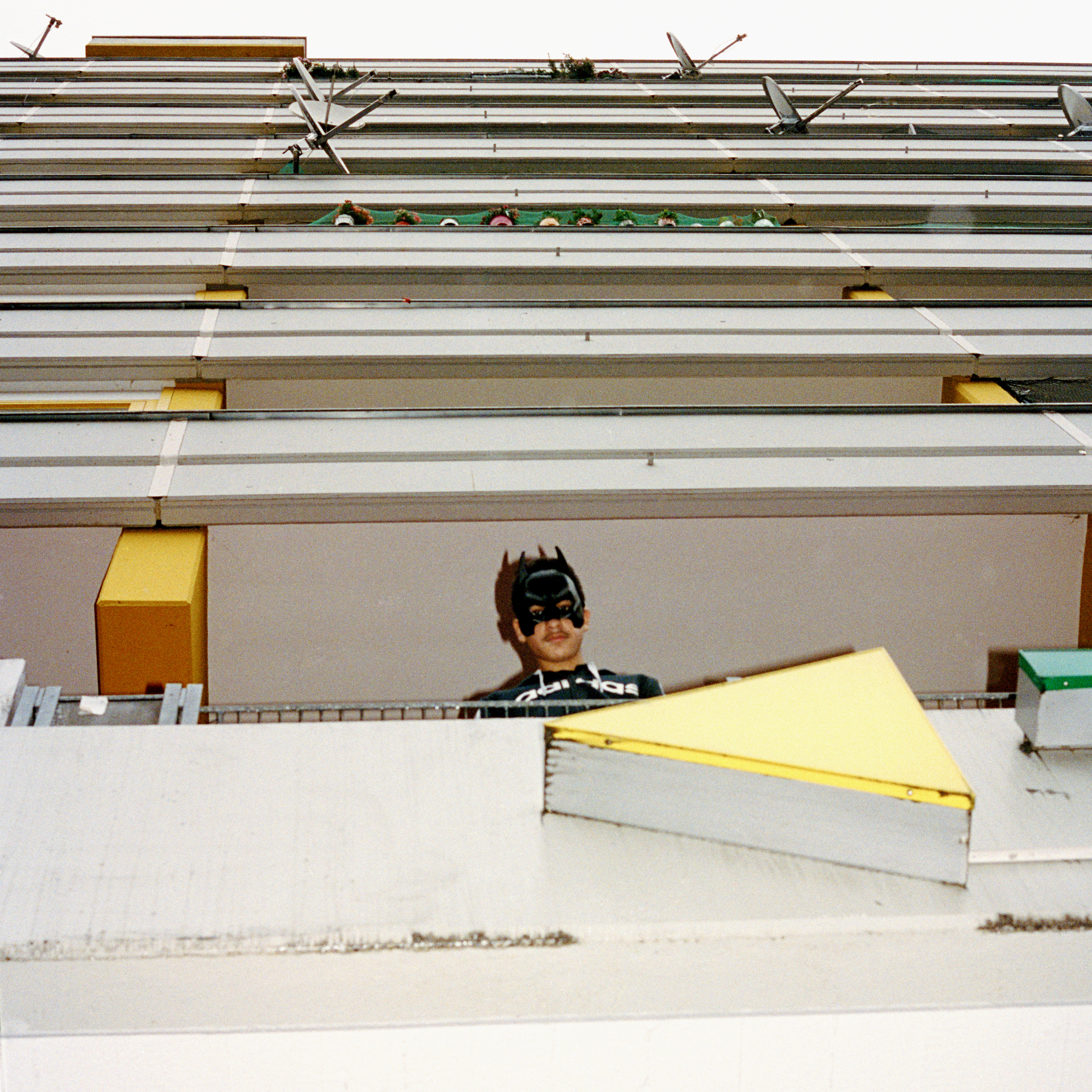 Kottbusser Tor, Berlin. A teenager wearing aBatman mask looks down from a white balcony with colourful shapes.