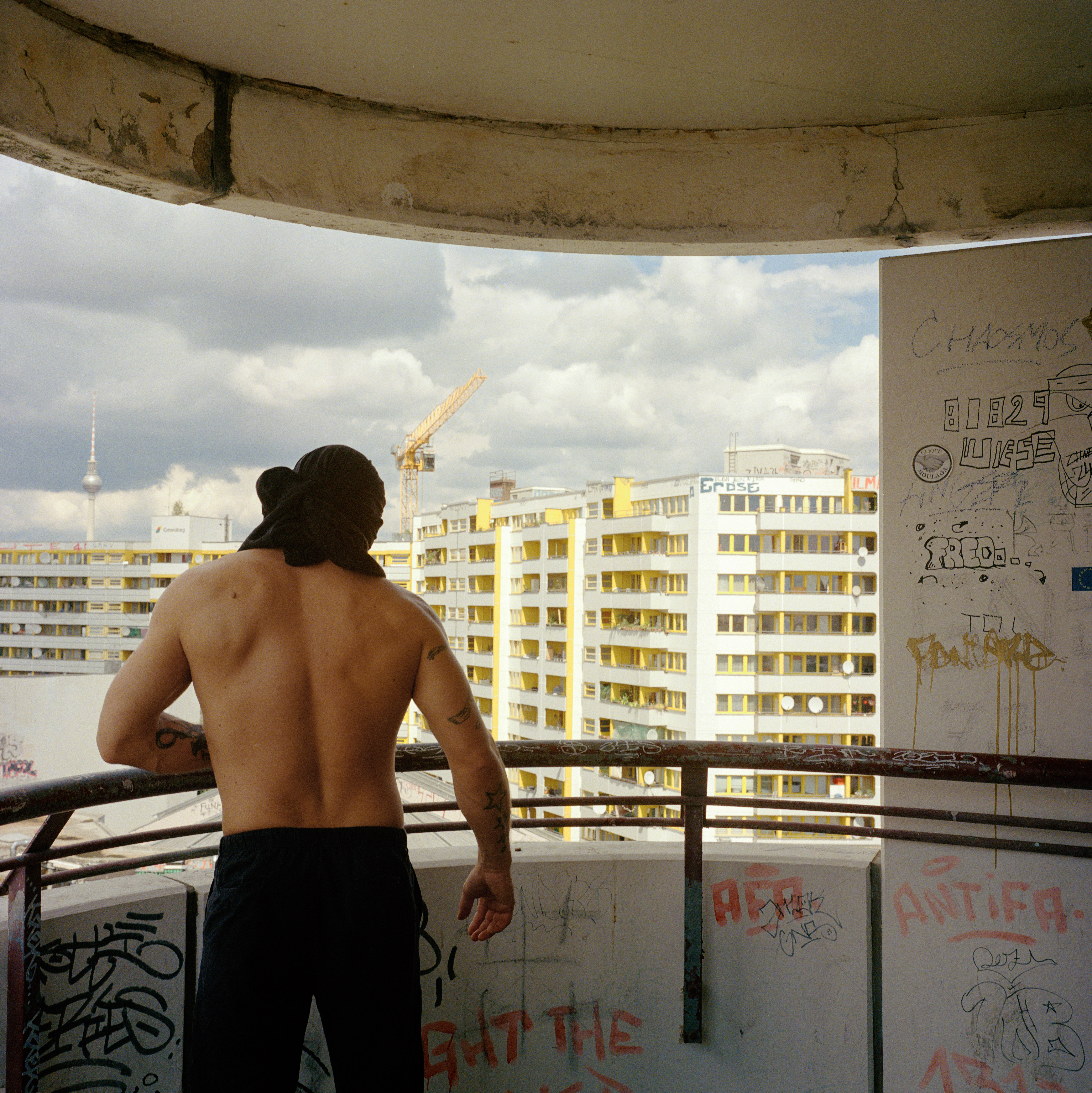 Kottbusser Tor, Berlin. A topless man with black pants and a black t-shirt on his head looks out from the balcony towards high white and yellow buildings under construction, the Berlin TV tower and a crane. There are graffitis on the balcony.