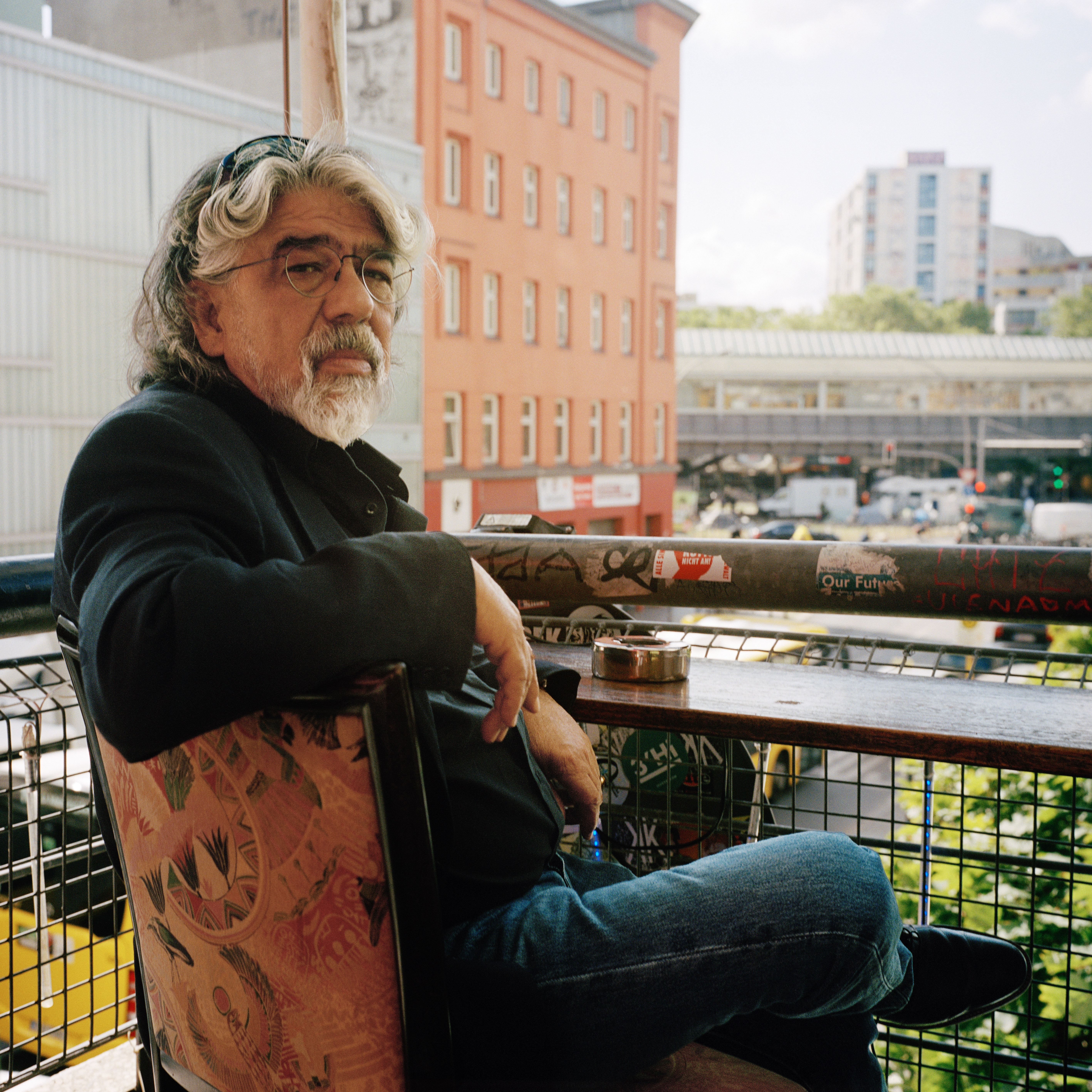 Kottbusser Tor, Berlin. A man with grey hair and a beard sitting on a chair in front of a balconly table, there's an ashtray on the table and some buildings and street lights in the background. He's wearing jeans, black boots, a black jacket and glasses.