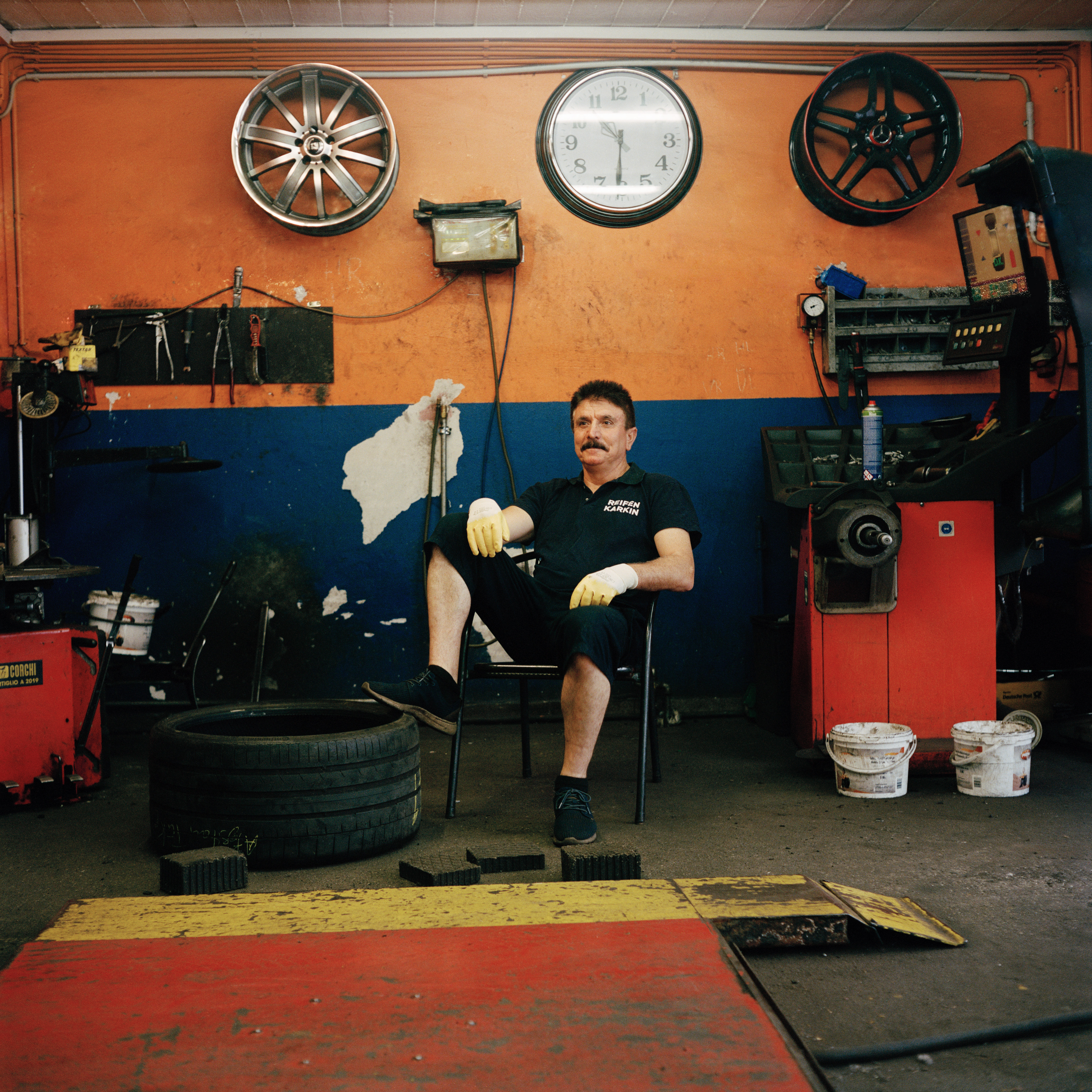 Kottbusser Tor, Berlin. A man sitting in a tyre shop. He has dark har, wears black shorts and a t-shirt and white gloves. His foot is on a big tyre. The wall in the background is orange and blue and there's a big watch and two wheel rims on it.