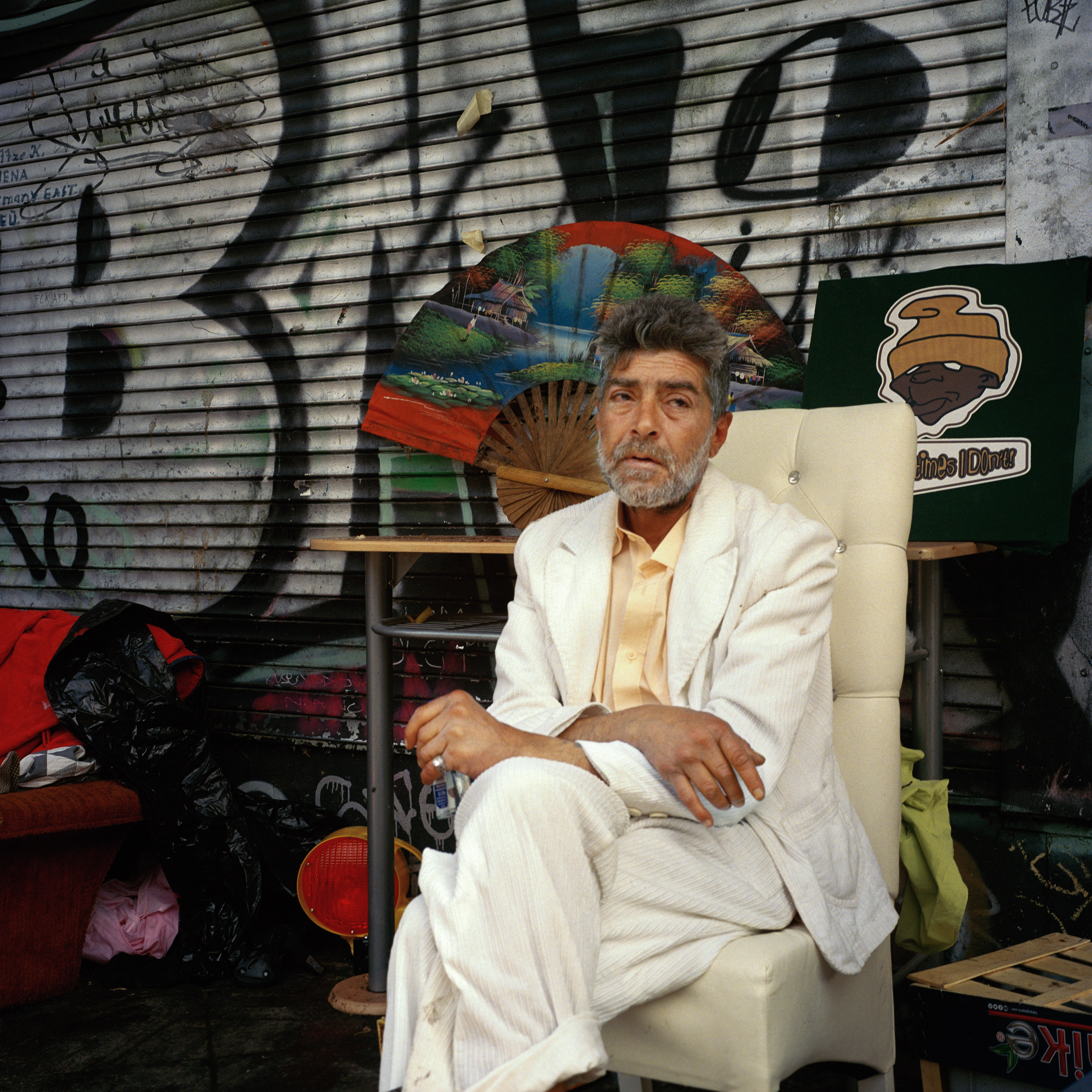 Kottbusser Tor, Berlin. An old man with grey hair and a beard in a white suit sitting on a white chair. In the background: random objects, a garage door with graffitis and a big colourful fan.