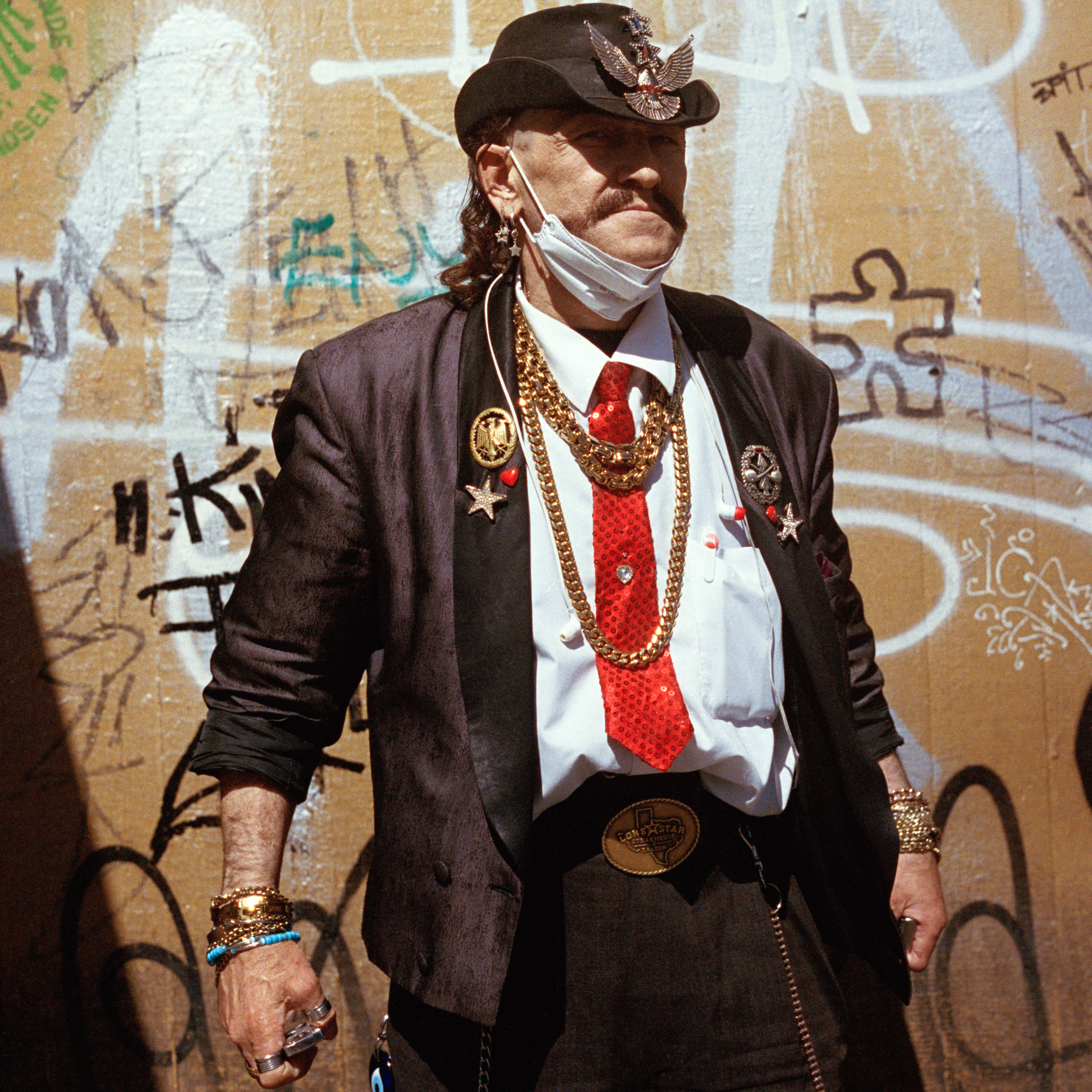 Kottbusser Tor, Berlin. A man wearing a brown cowboy hat and jacket with golden decorations. He also wears a white shirt and a red tie as well as golden necklaces. He has long hair.