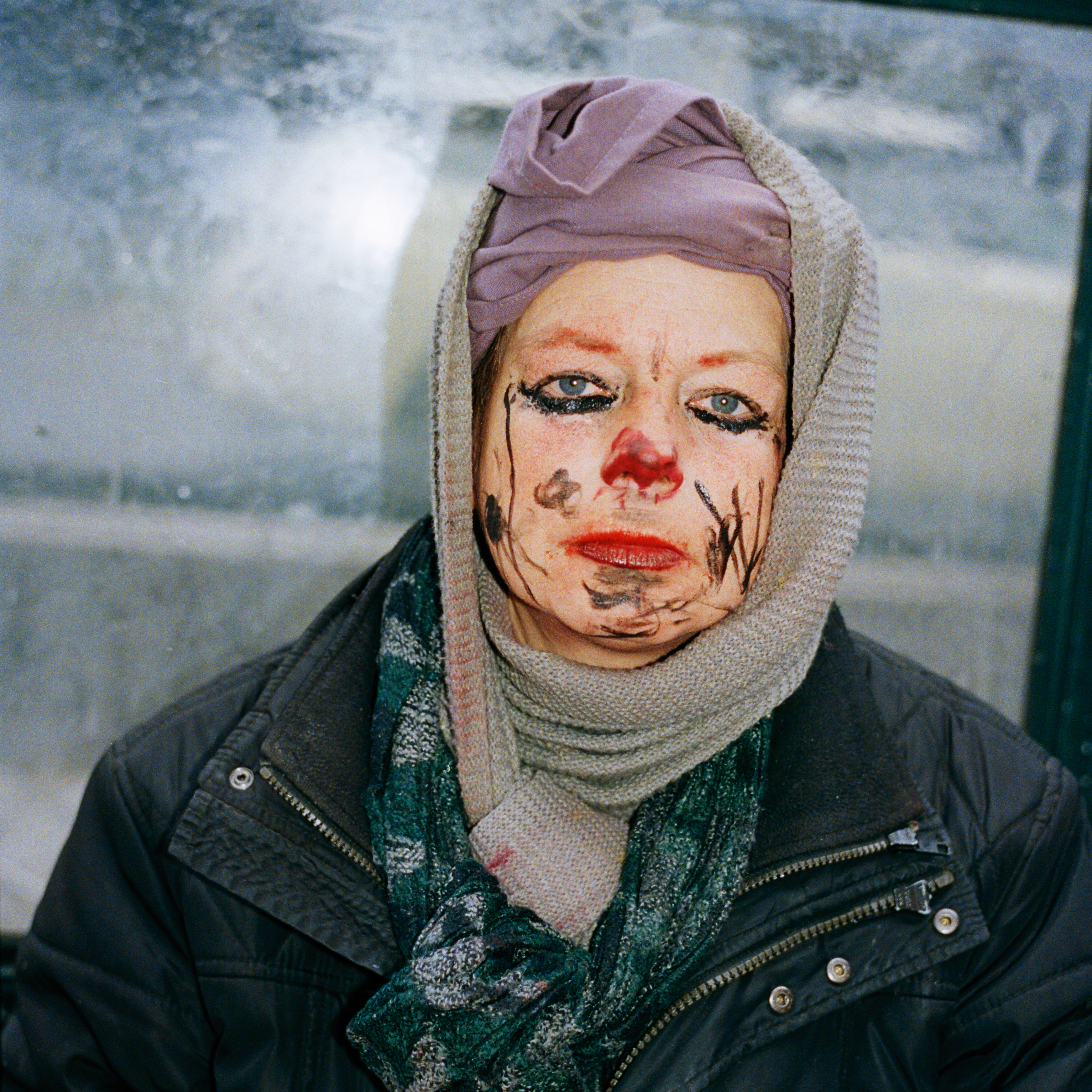 Kottbusser Tor, Berlin. A woman with black and red makeup and drawings on her face. She's wearing purple material and a grey scarf on her head, a green scarf around her neck and a black jacket.