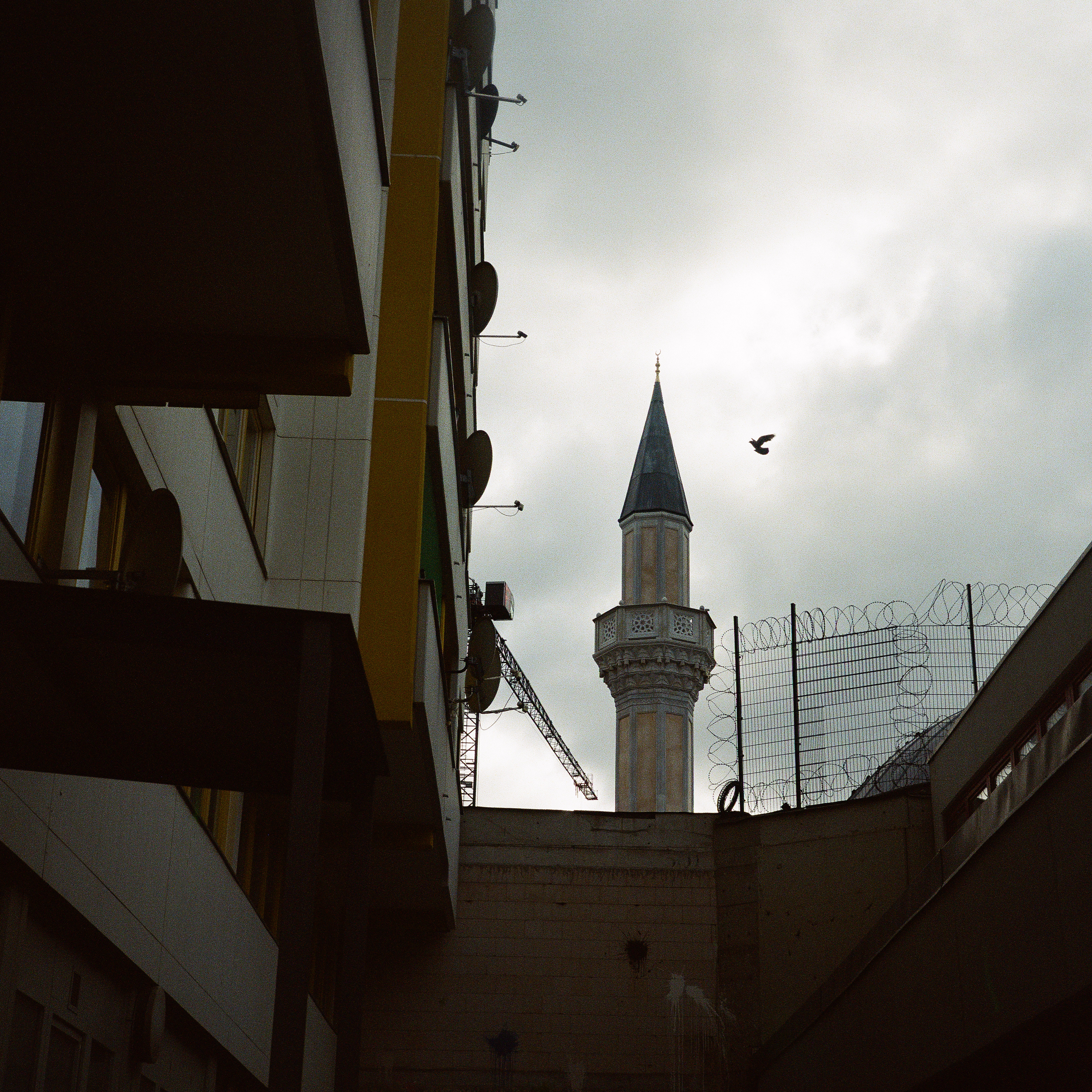 Kottbusser Tor, Berlin. A backyard with a view on a mosque and windows of a building. There's barbed wire and a bird in the grey sky.