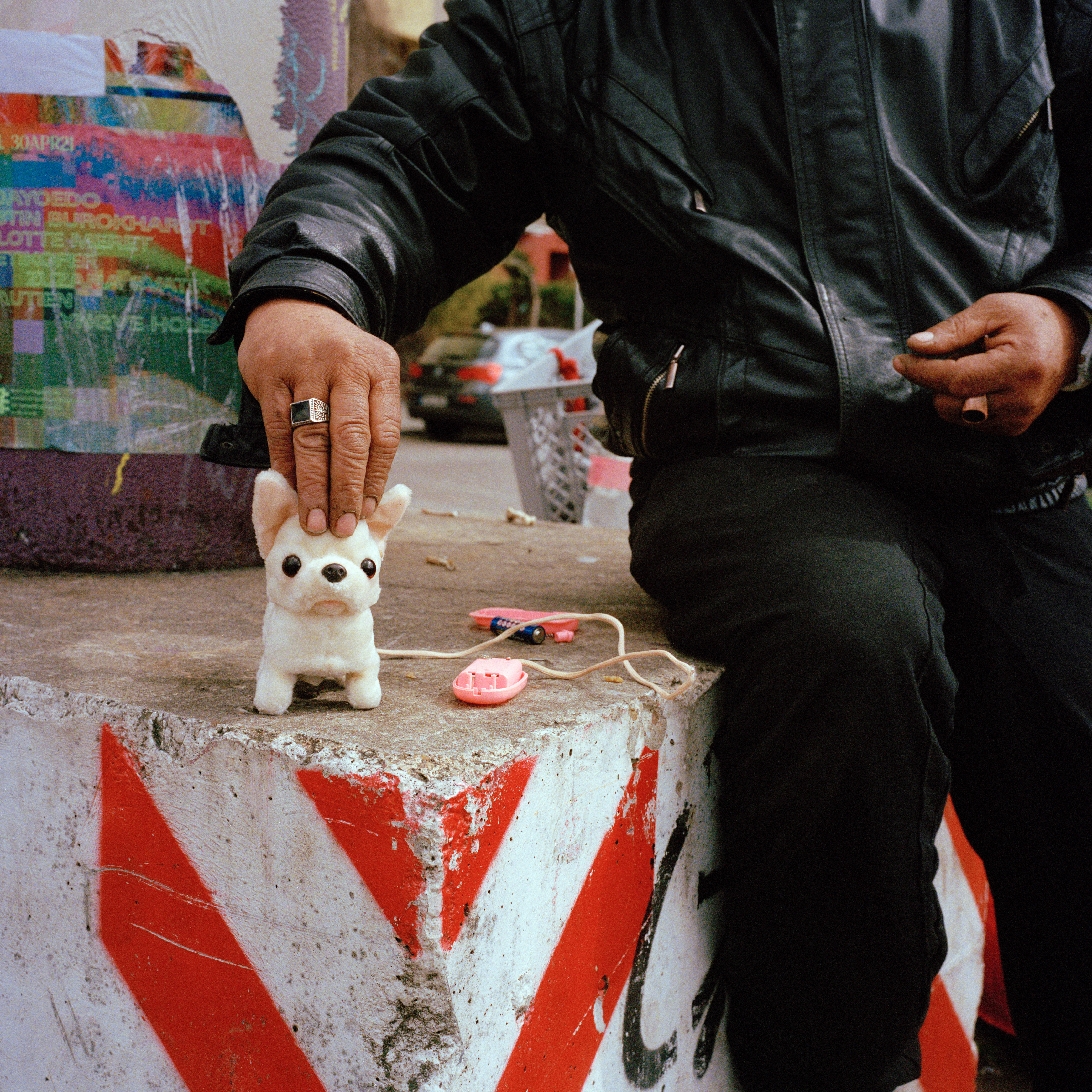 Kottbusser Tor, Berlin. A dog game with a pink handle. A man wearing black sits on a wall and holds the game's head. There's a colourful poster in the background.