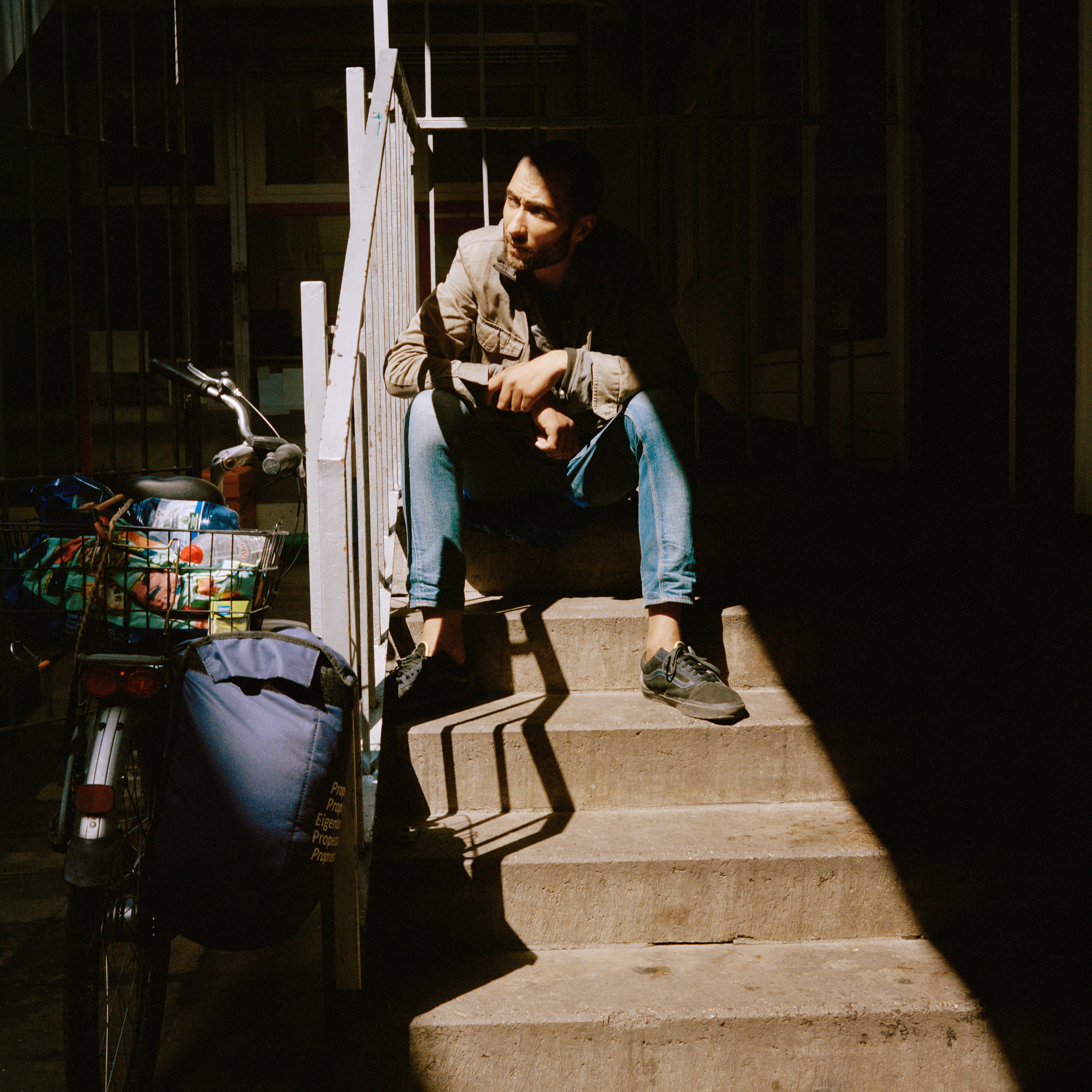 Kottbusser Tor, Berlin. A man sits on a partly sunny staircase, looking towards the light. He's wearing blue jeans, a kaki jacket and black shoes. There's a bike with a filled basket and a bag nest to the stairs.