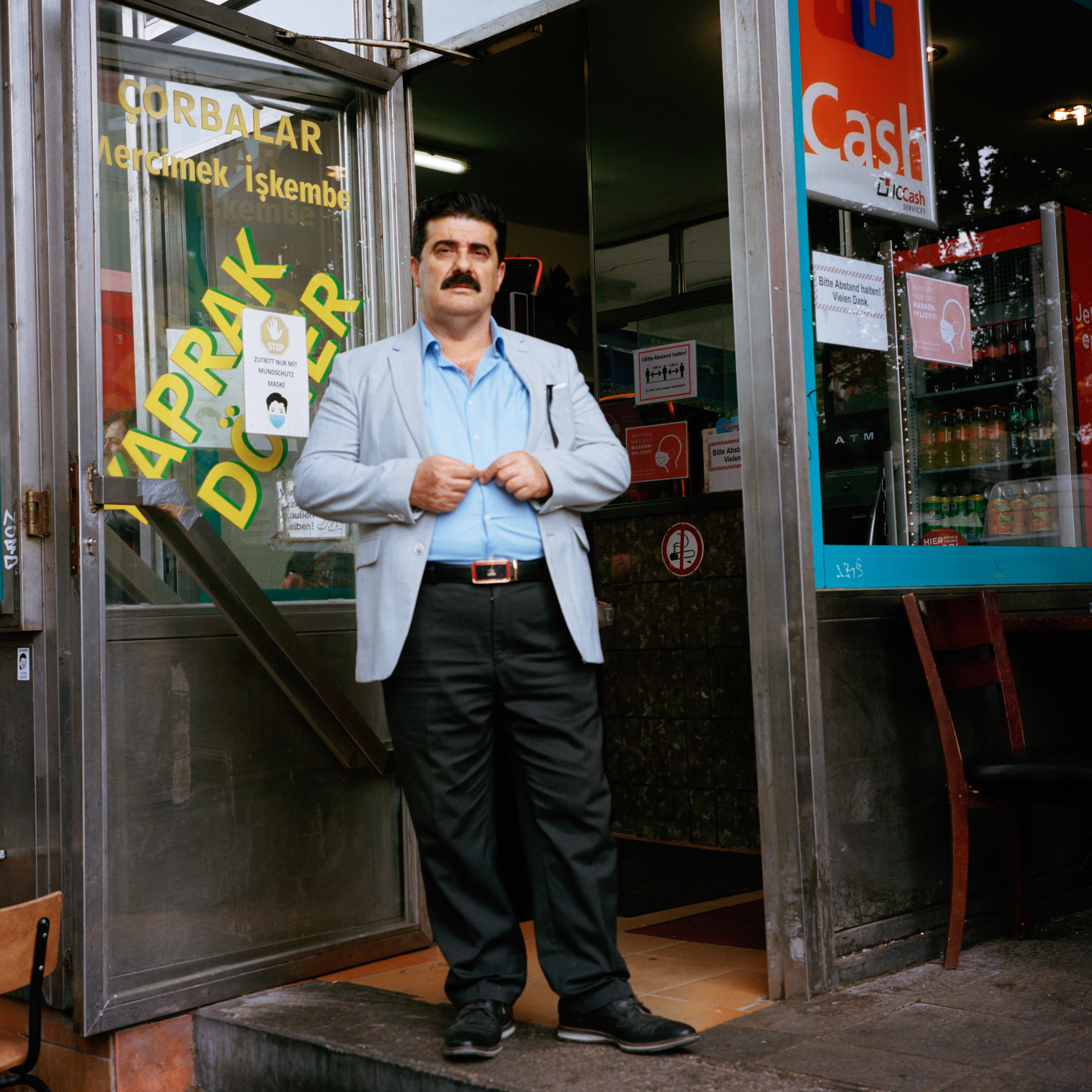 Kottbusser Tor, Berlin. A man wearing a sky-blue jacket and shirt, black pants and shoes, stands in the entrance of a kebab.