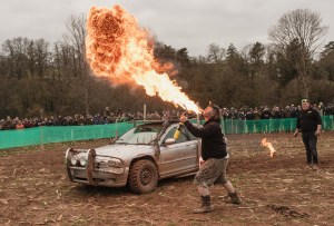 A man spews a huge ball of flame from his mouth, he’s holding a propane torch and is standing in an open dirt field with spectators in the distance. A modified off-road sedan is behind him.