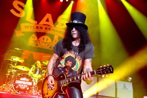 A photo of slash from Guns N Roses b and playing guitar in a black tophat on stage.