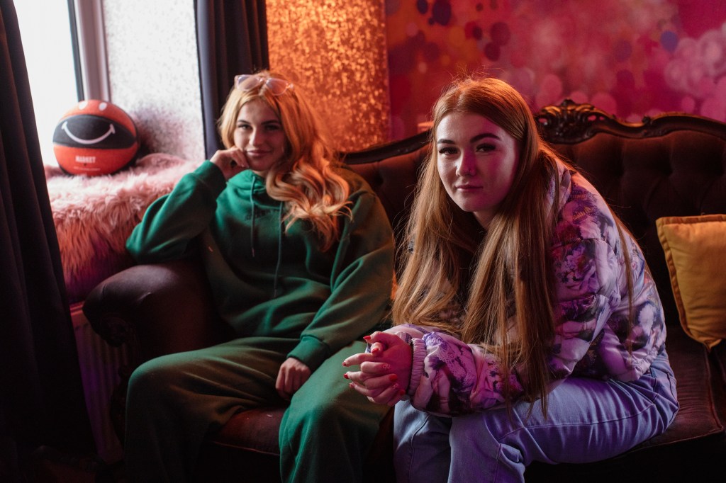 Sophie Aspin and Millie B sit on an ornate couch inside a warmly lit room. Sophie wears a green sweatsuit, Millie B wears a purple puffy jacket.