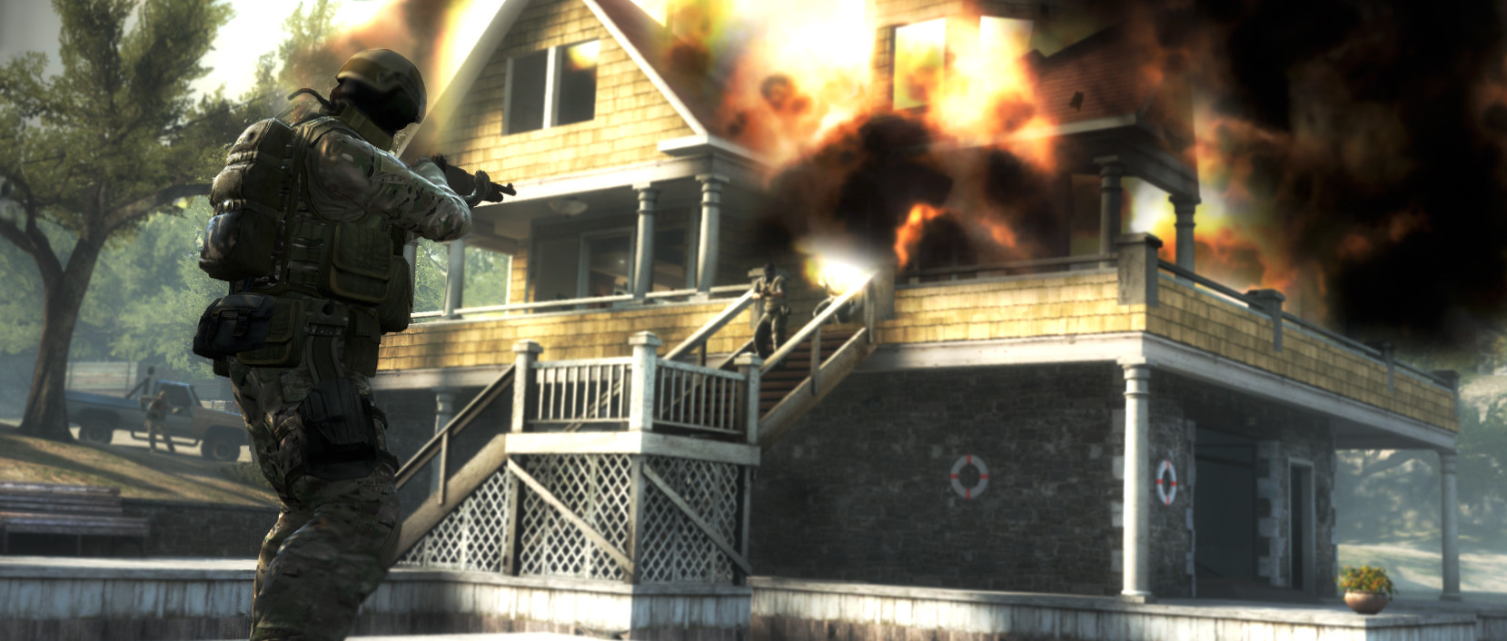 A soldier wearing tactical gear aims a shotgun at a man standing on the external stairway of a building, which is engulfed in a fireball as something inside explodes.