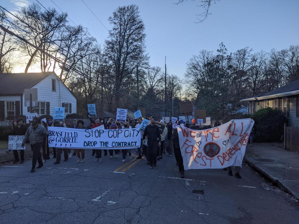 Protesters march down a street in Atlanta holding banners that say "Stop Cop City / Drop The Contract" and "We Are All Forest Defenders"