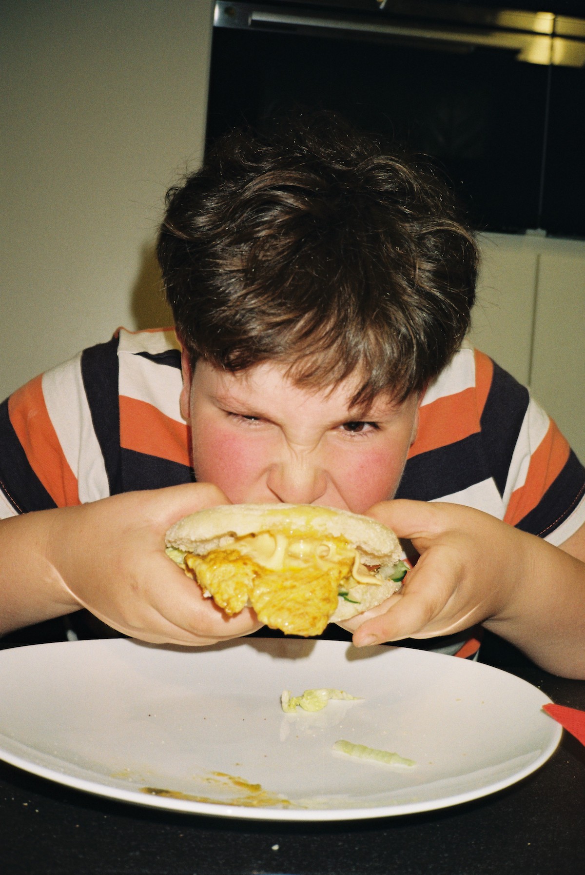 Ramadan photos. A kid wearing a black, orange and white stripe shirt biting on a juicy burger.