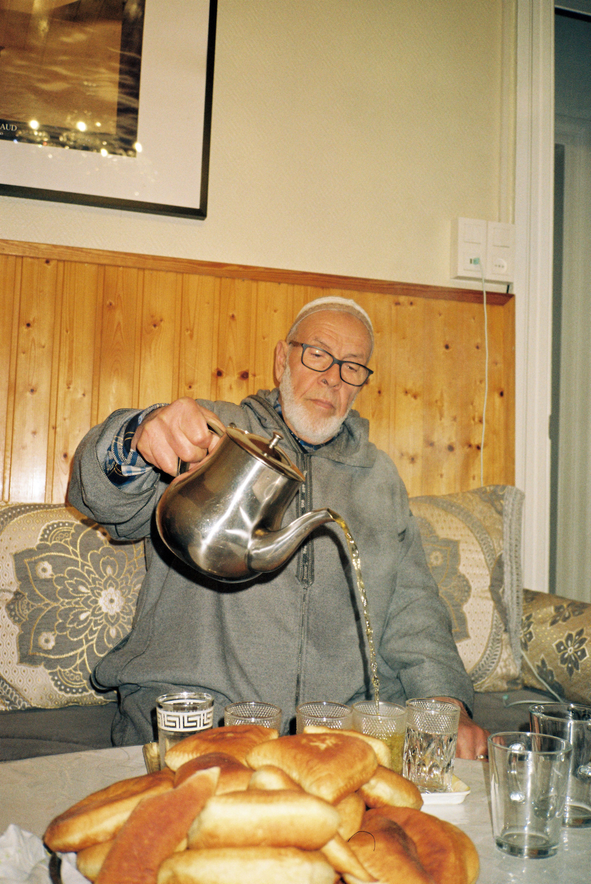 Ramadan photos. An old man wearing a grey sweater and sitting on a shiny couch, he's serving mint thee in glasses and there's a pile of pastries on the table.