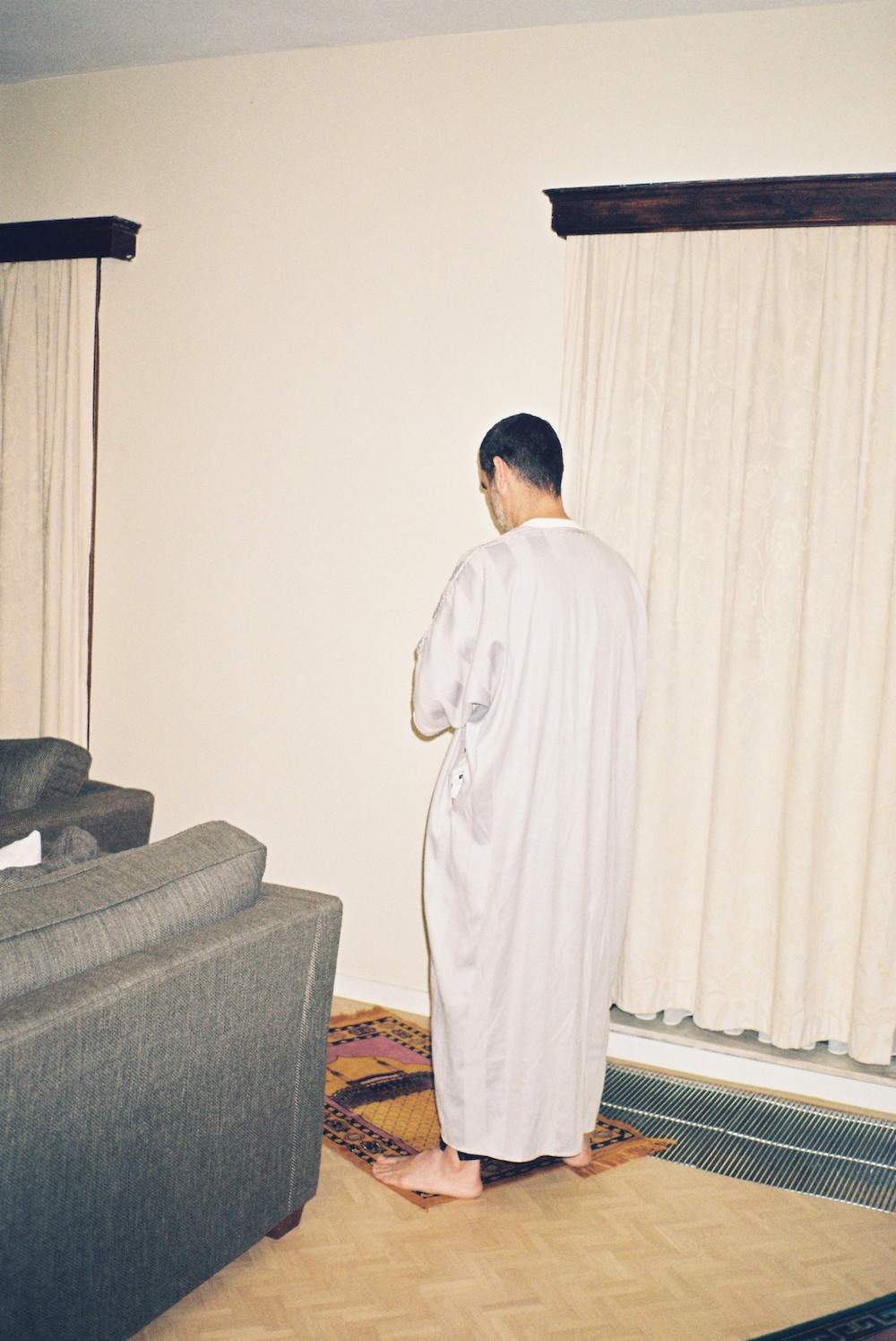 Ramadan photos. A man praying on an orange carpet next to a grey couch. He's wearing a white jellaba and there's white curtains in the background.