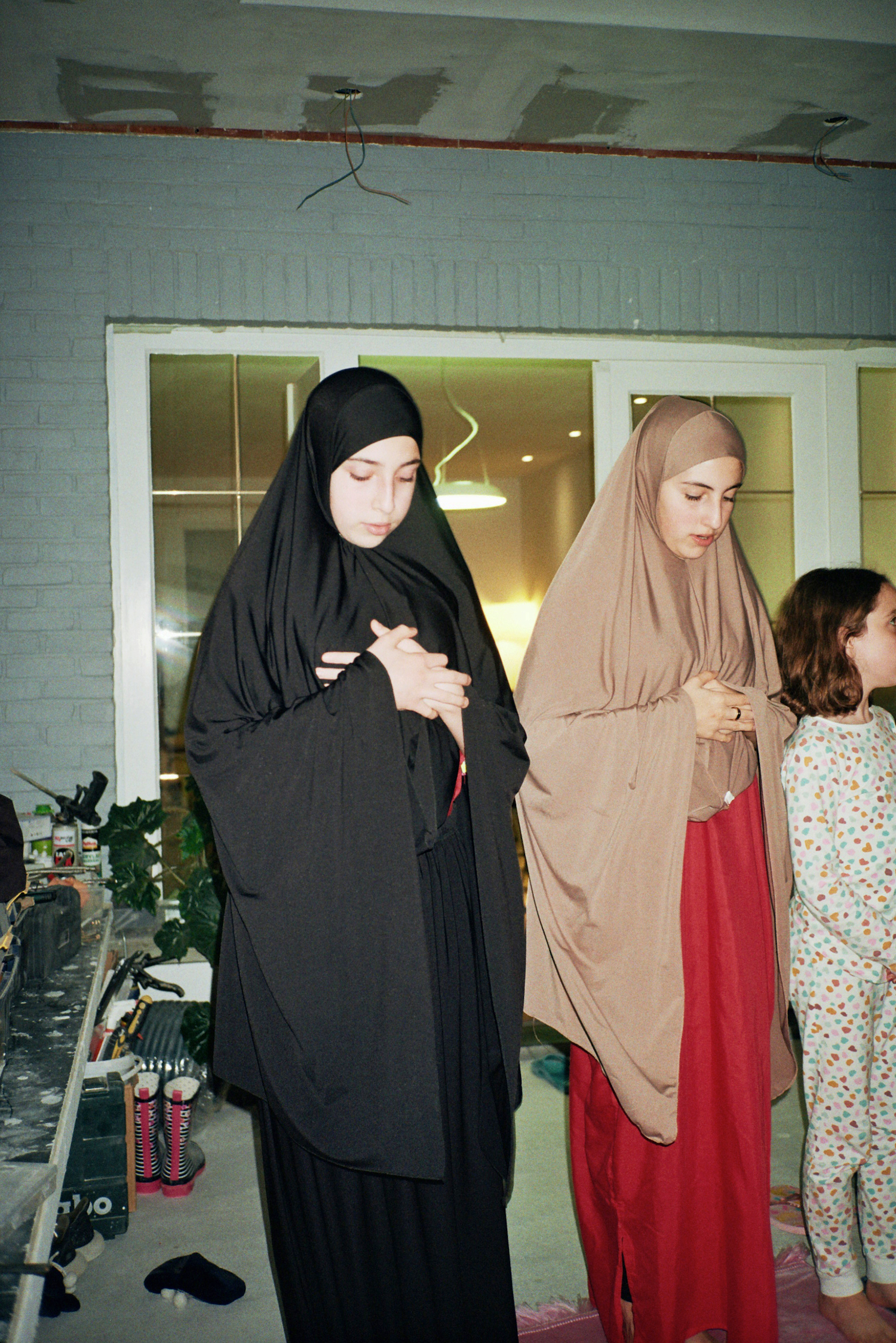 Ramadan photos. Two women and a kid praying. One is wearing a black hijab and long dress, the second one is wearing a red dress an a beige hijbab, and the kid is wearing colourful pyjamas.