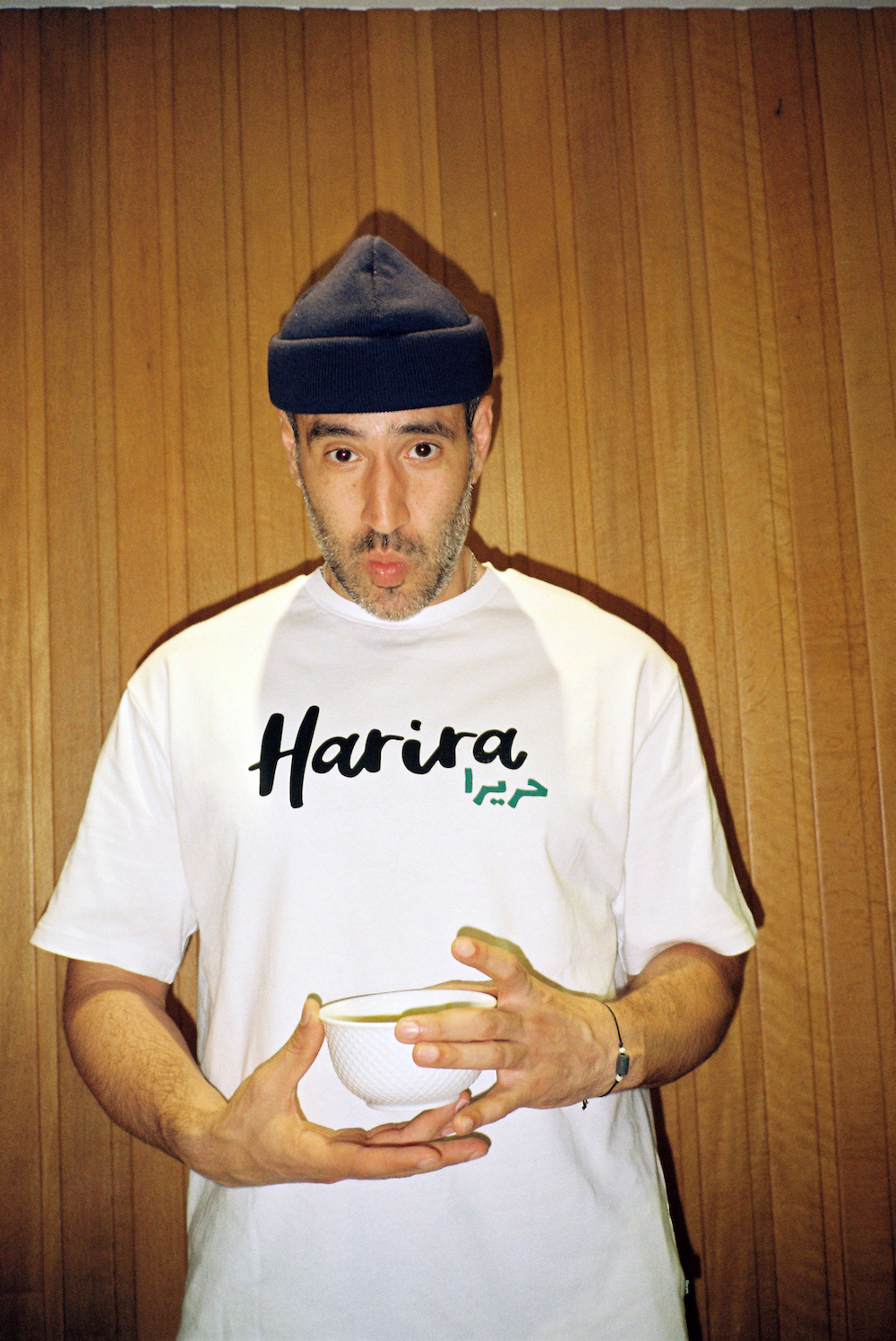 Ramadan photos. wearing a white shirt with harira written on it and a black beanie is holding a bowl of soup.