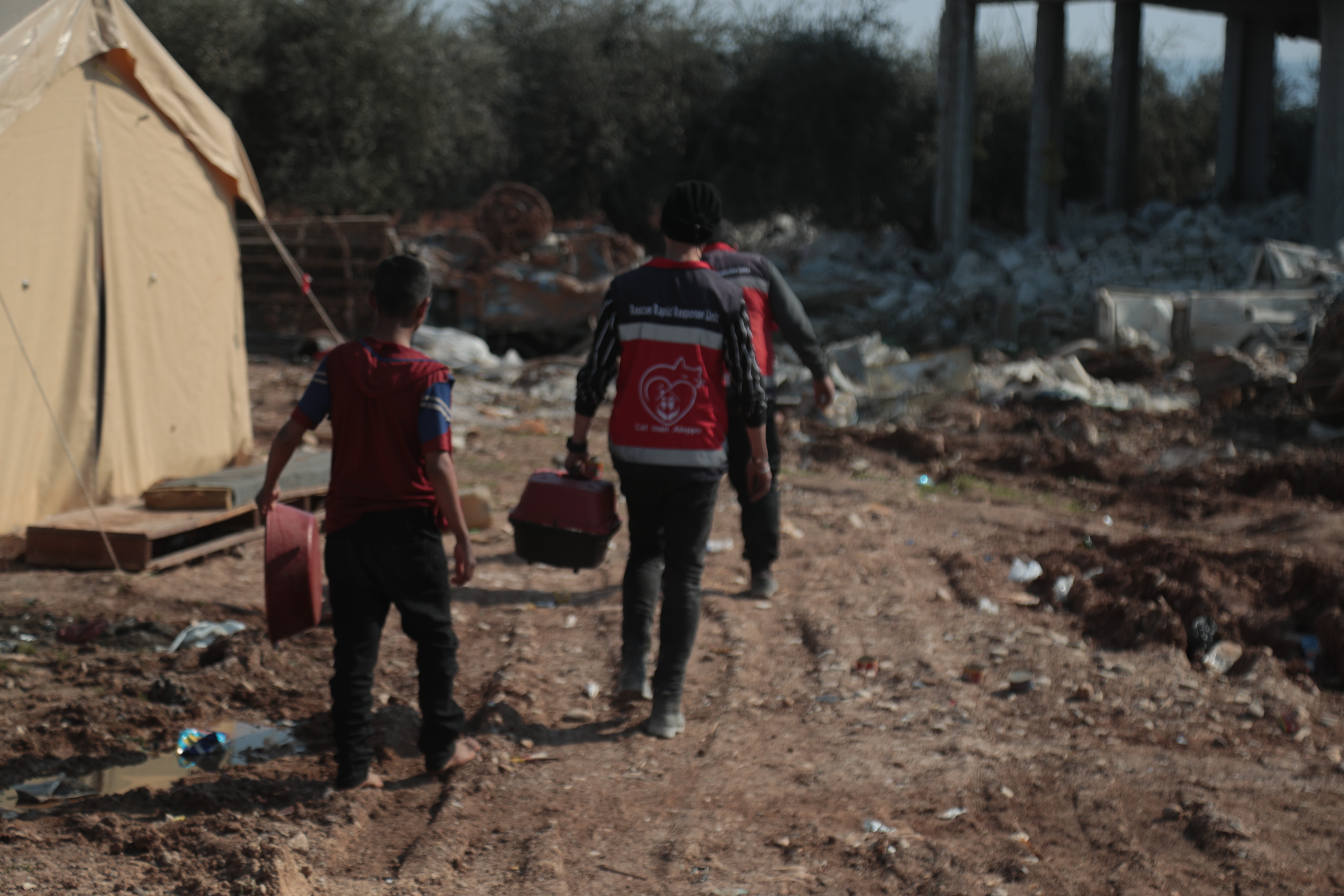 Mohammed Alaa Aljaleel, Cat Man of Aleppo – two men and a child carrying a cat carrier and a red basin and walking in an open space filled with rubble near a tent