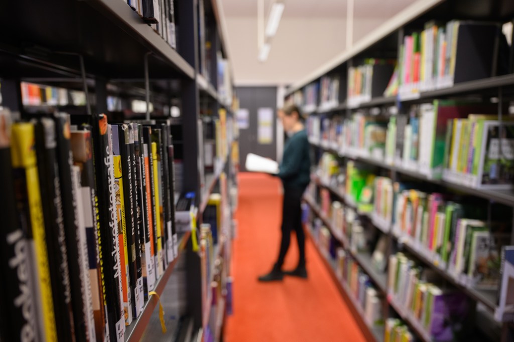 A person stands reading a book in a library surrounded by shelves of books