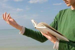 A person reading from a book of poetry with an ocean in the background