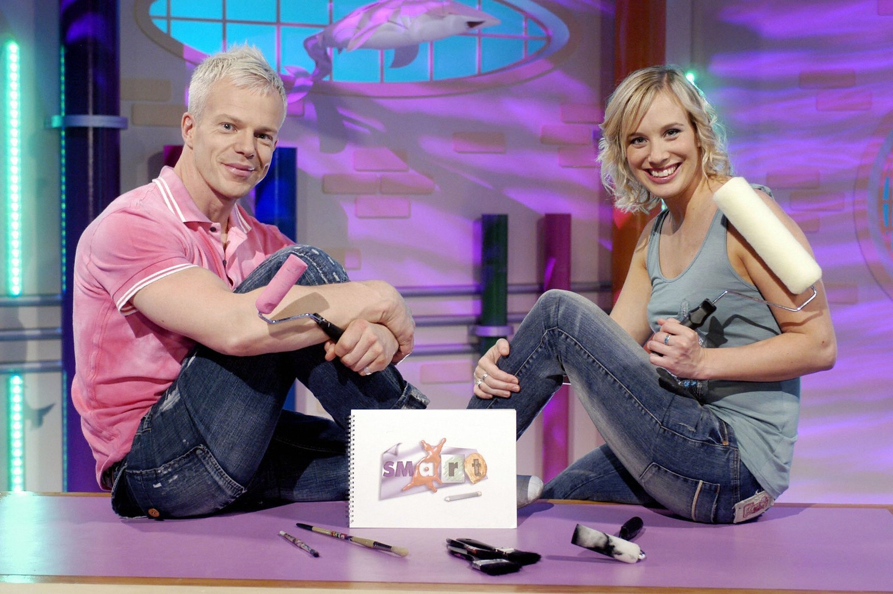 Mark Speight and Kirsten O'Brien presenting SMart, sitting on a table holding paint rollers behind an art pad, pens and brushes.