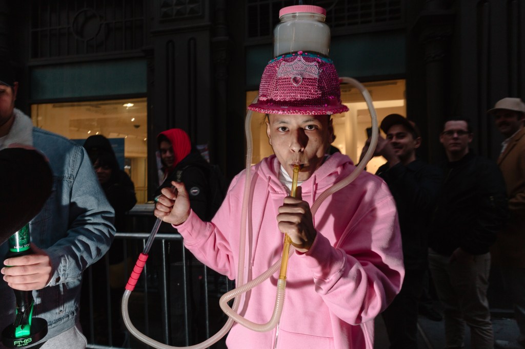 A man in a pink hoodie smokes out of a tube that is connected to his glitter hat that has a smokingto