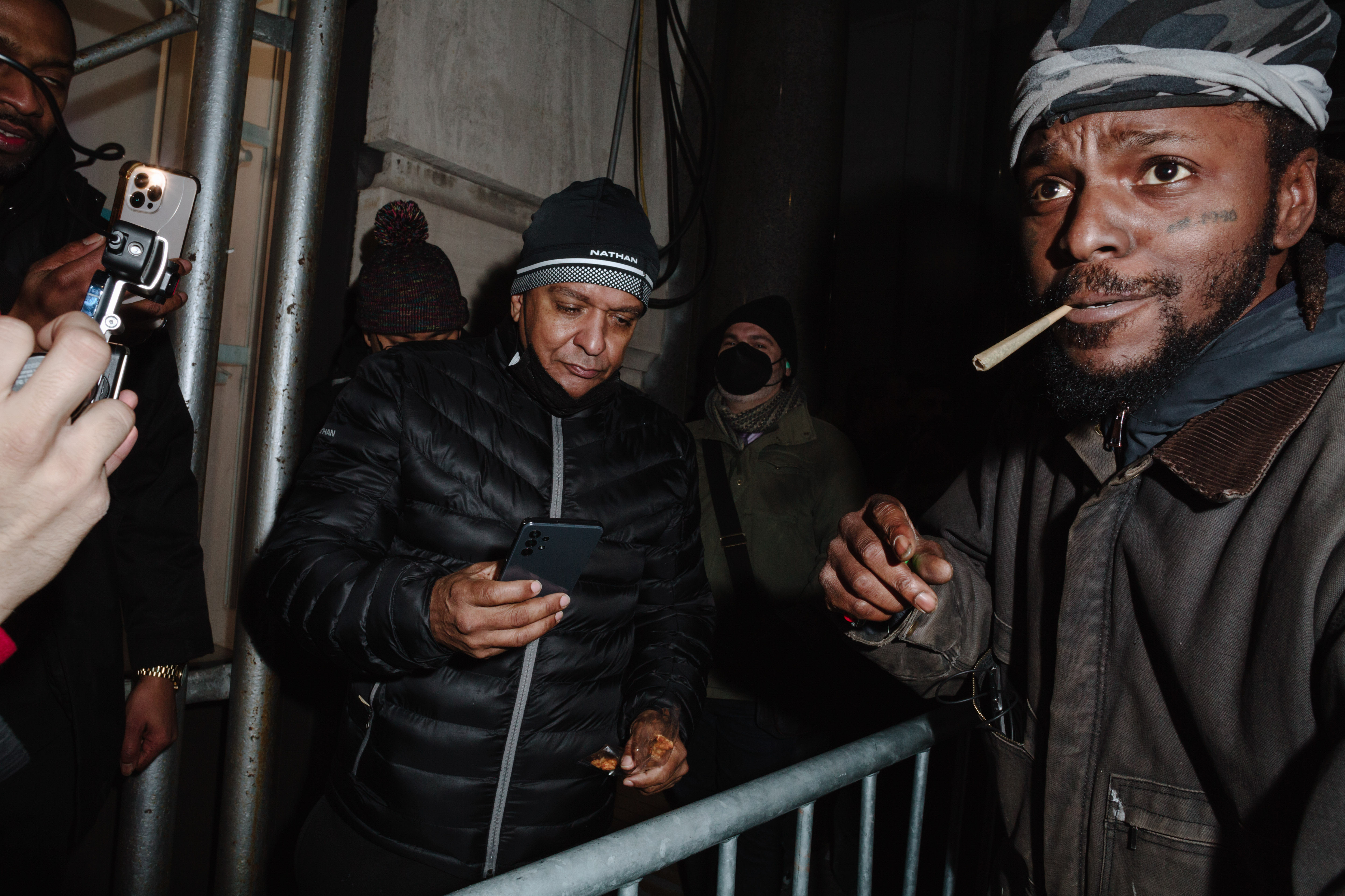 A man holds an unlit joint in his mouth, another man is seen on his phone behind a metal barrier.