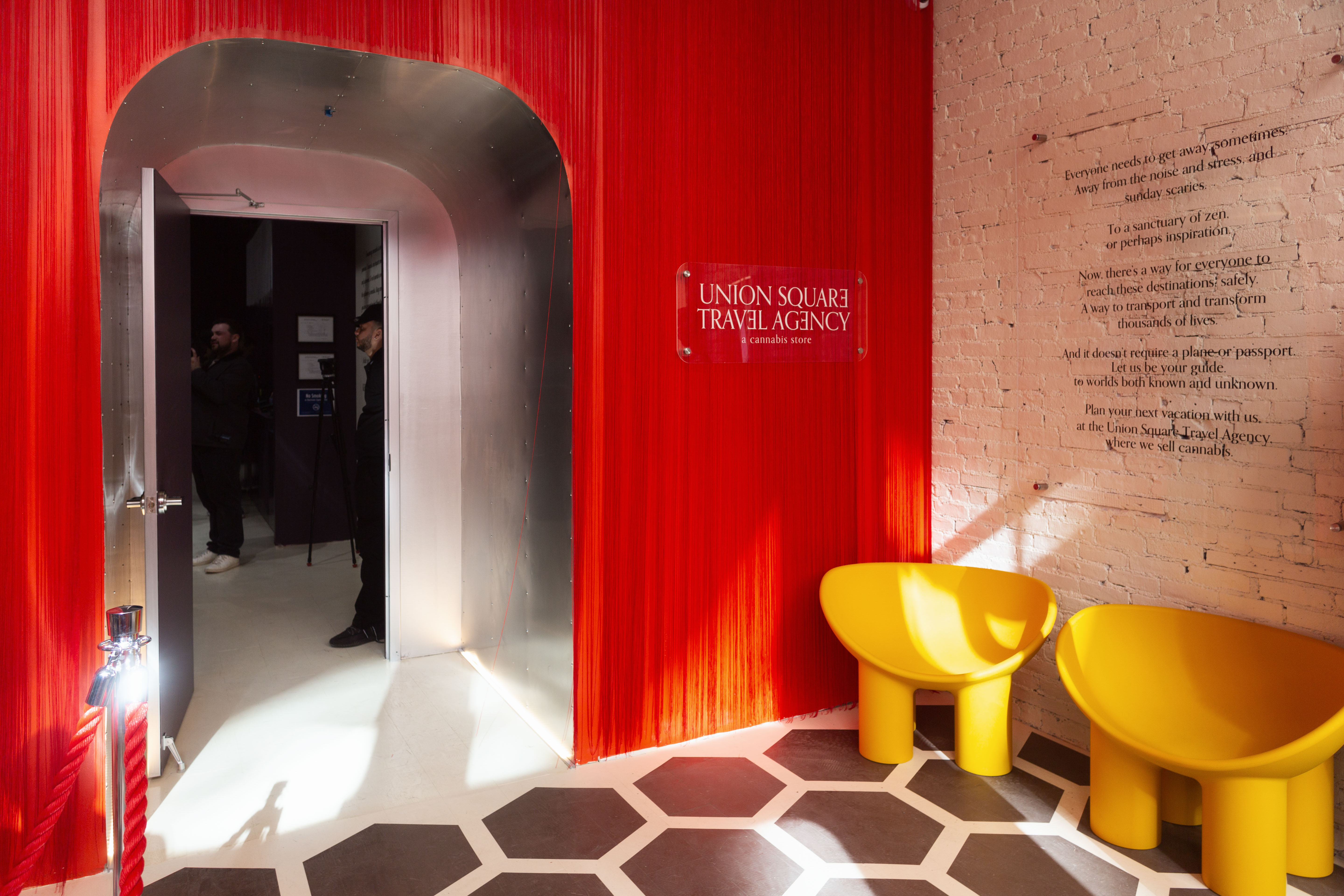 A fancy storefront with a red wall and modern yellow chairs, red braided rope dividers can be seen in the corner, and the storefront name is printed on a clear placard that reads Union Square Travel Agency.