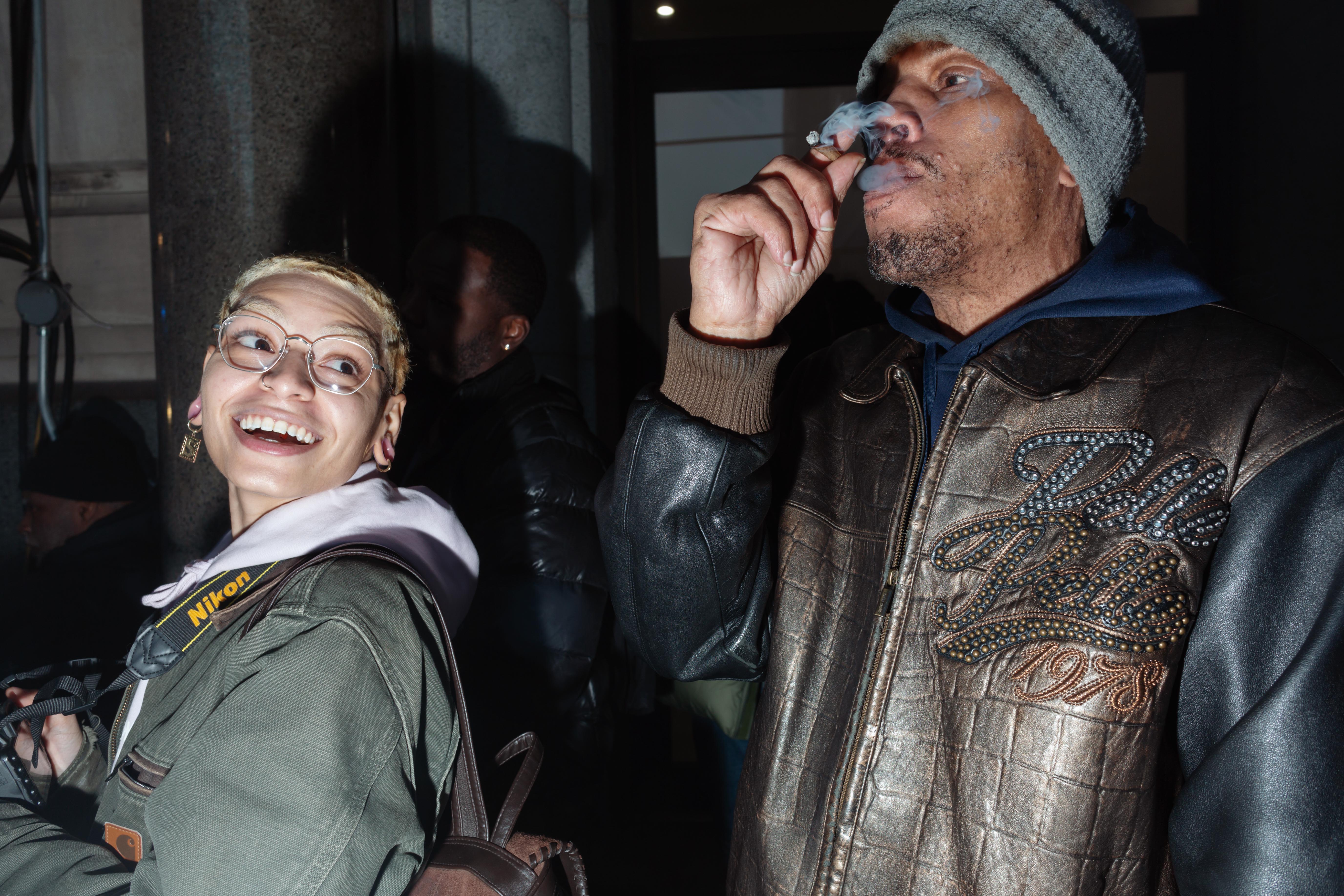 A man takes a puff of a blunt while a woman with a camera looks behind her shoulder towards him.