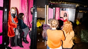 Left: three women inside a small pink trailer. One is getting naked, one is looking at her, the third is laughing and looking at someone off camera. Right: brunette in red lingerie stripping inside a black and pink trailer in front of a small crowd.