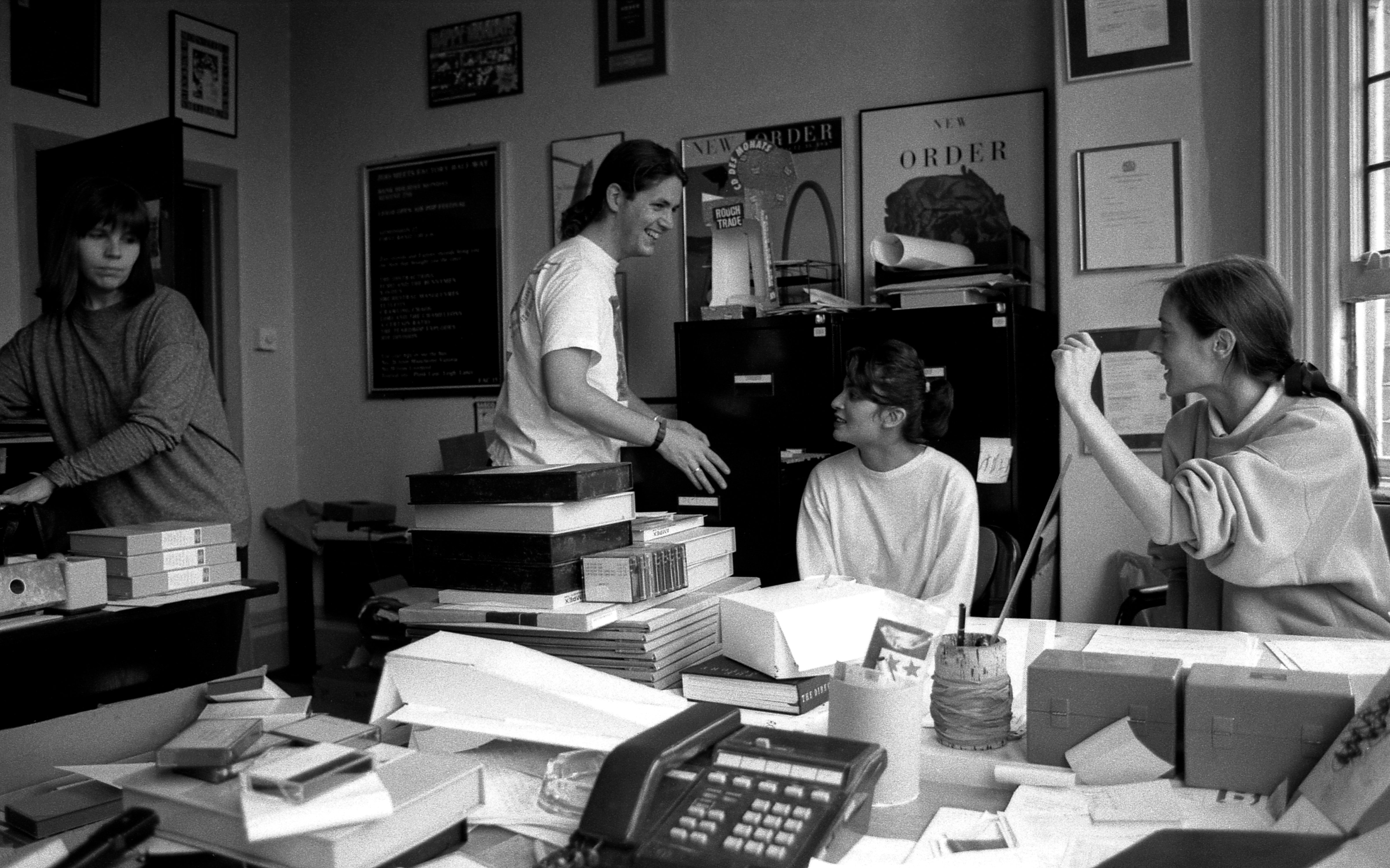 Four women chat with each other in an office space, the desks are piled high with paper, negative binders, and books.