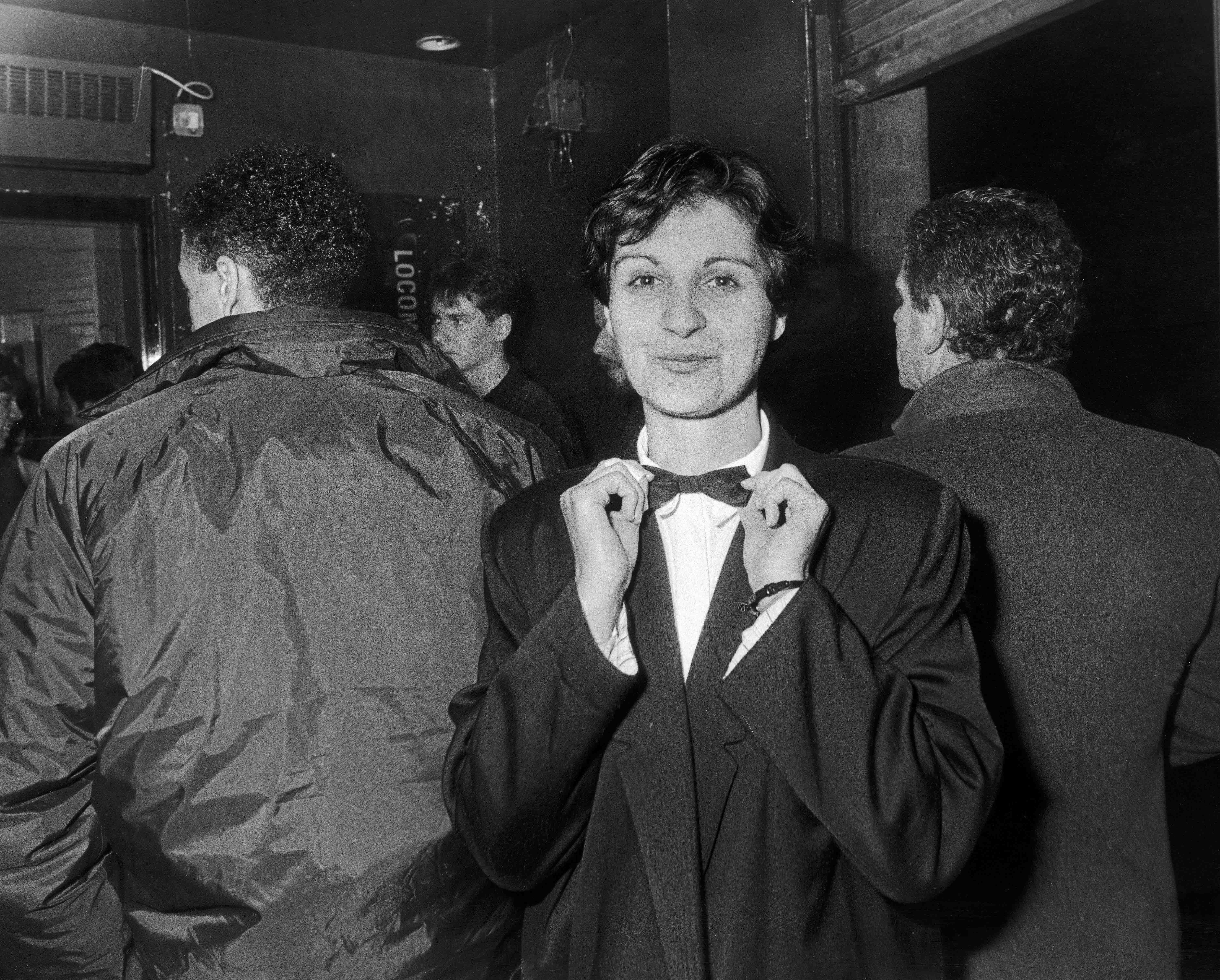 Yasmine Lakhaney is pictured wearing a suit and a bowtie looking at the camera, behind her are two mens' backs.