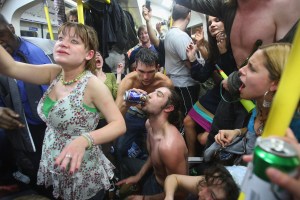 A photo of chaos on a tube carriage - a man pouting beer into his topless friends mouth, a topless man with nipple tassles standing, a girl shouting, everyone dripping in sweat.