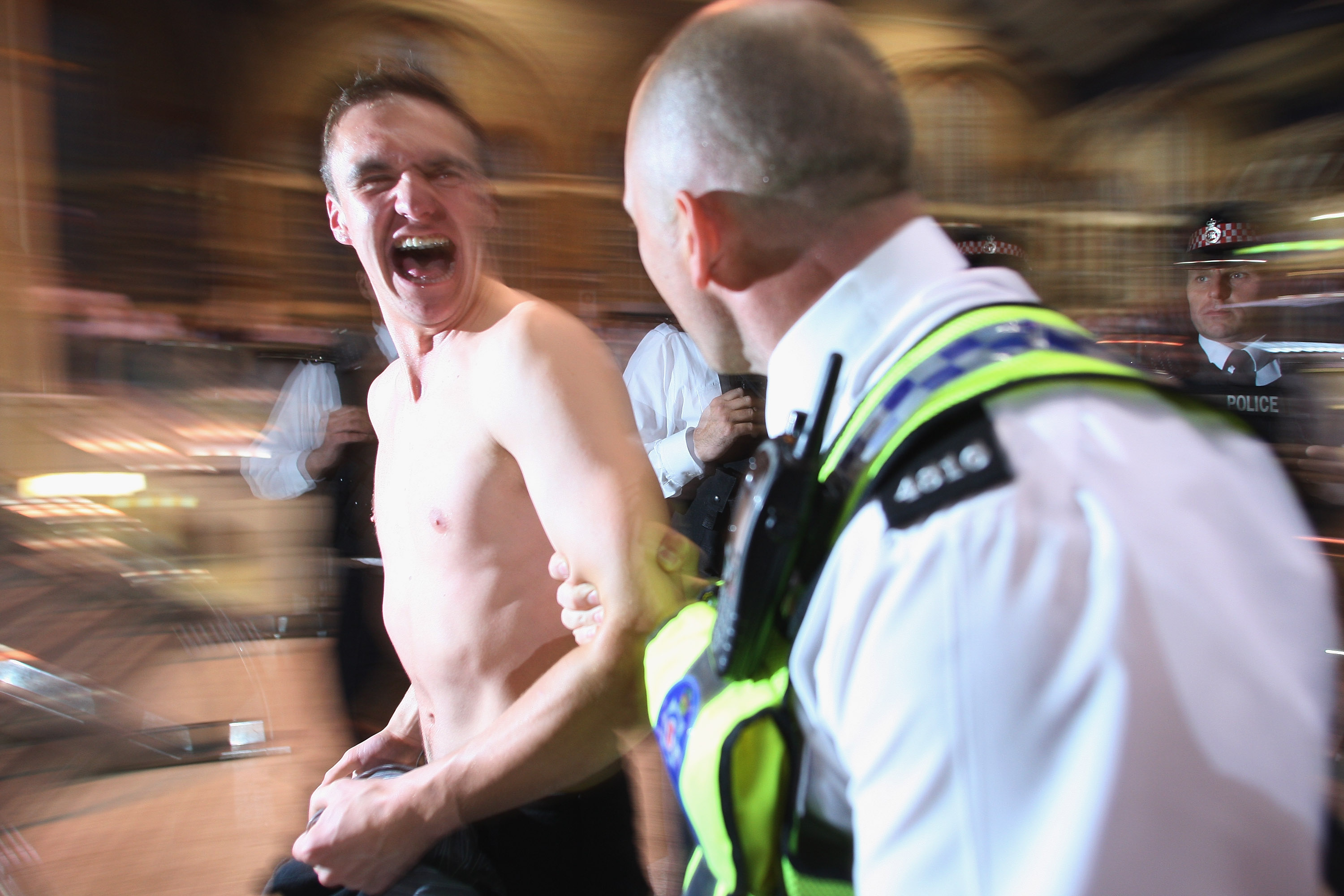 A photo of a topless man being escorted away by police officers in uniform, his face is laughing.