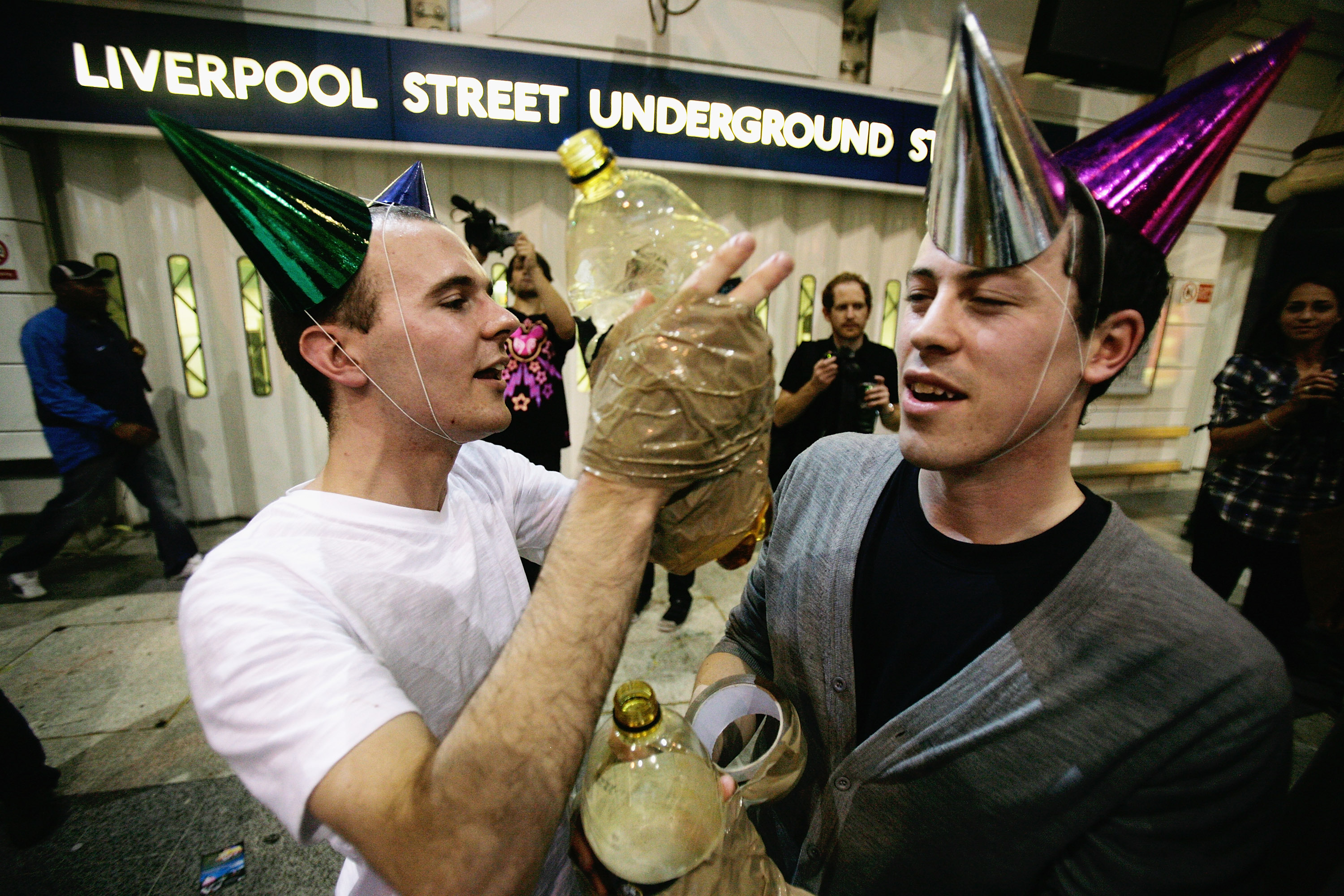 A photo of two drunk men standing outside Liverpool Street Station in party hats, with bottles of Strongbow cellotaped to their hands.