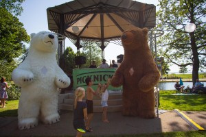 photo of three kids and two mascots, a white and a brown bear, standing in front of a kiosk with a sound system and DJs in park with a pond in the background.