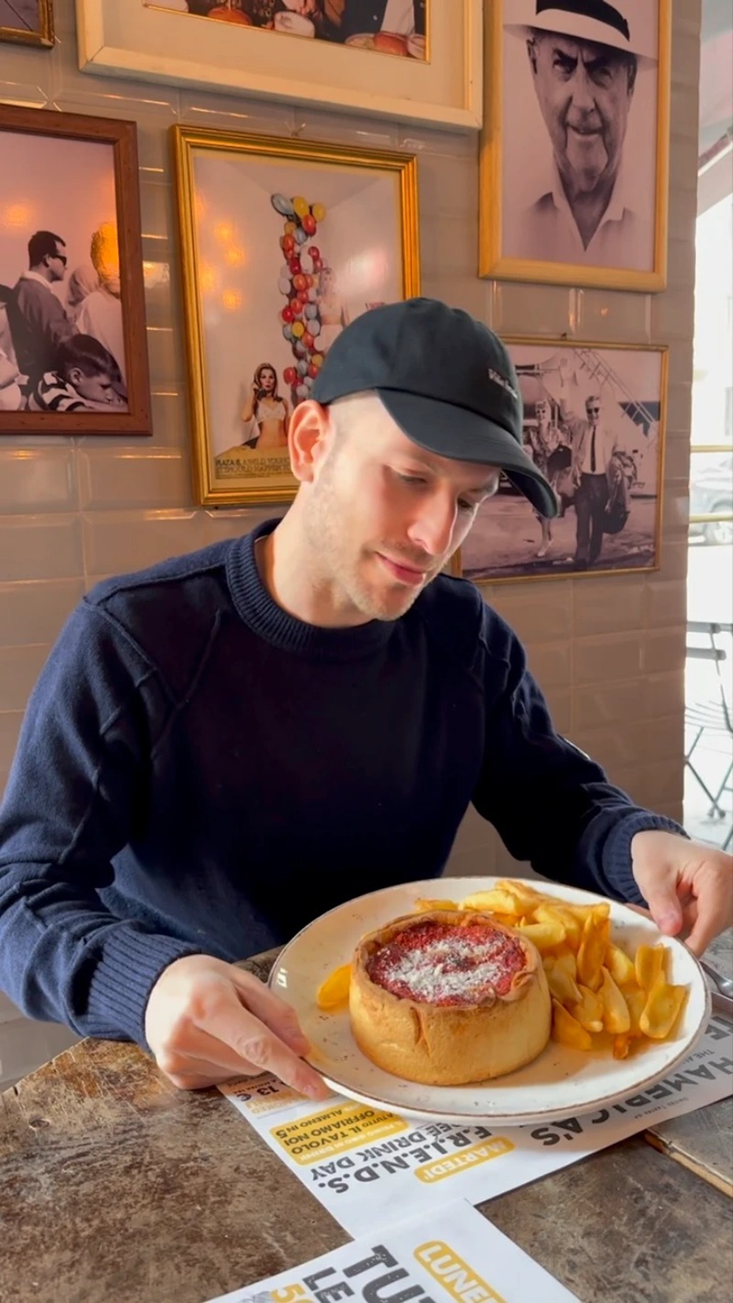 Chicago style deep dish pizza - photo of a man eating a pizza with fries. He's wearing a black cap and blue jumper and there are frames with photos in the background.