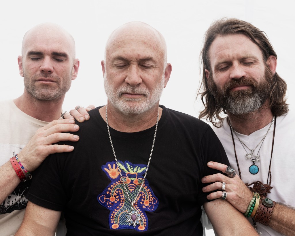 Een foto van de drie oprichters van de Church of Psilomexothin: Benjamin Moore, Ian Benouis en Greg Lake. Ze houden elkaar met gesloten ogen vast.