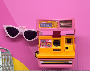 image shows polaroid camera and white sunglasses on a pink wall