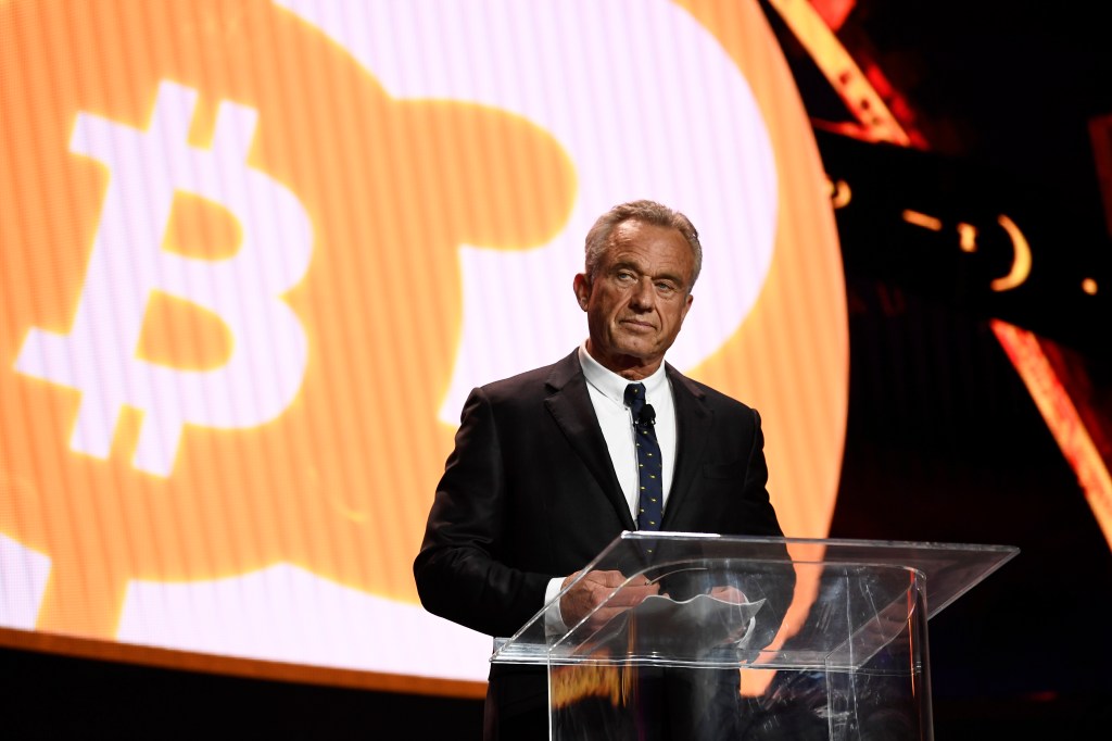 RFK Jr. at a glass lectern with an orange bitcoin symbol projec behind him